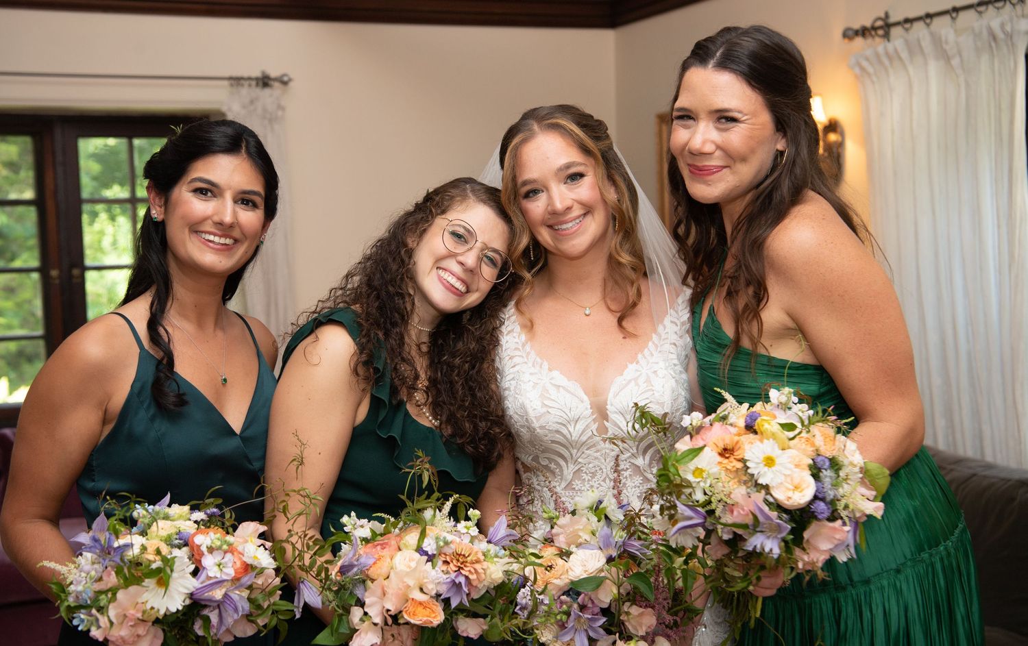 Bridesmaids in emerald green dresses holding colorful bouquets pose with bride in white lace gown.