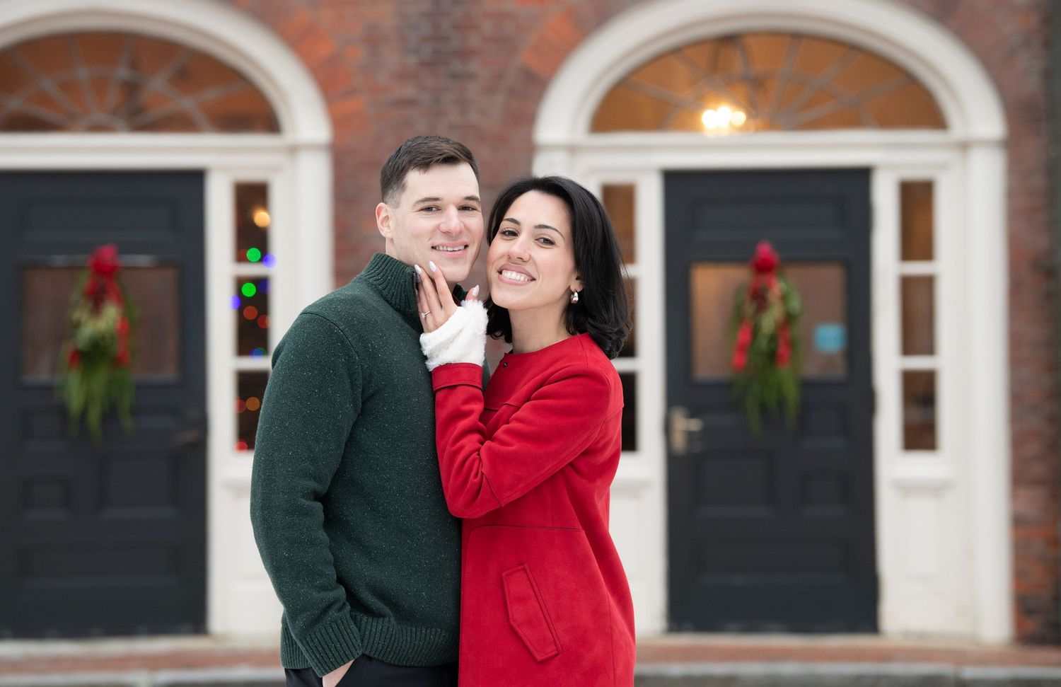 A couple in red and dark green winter outfits embrace and share a romantic moment outside a brick building.