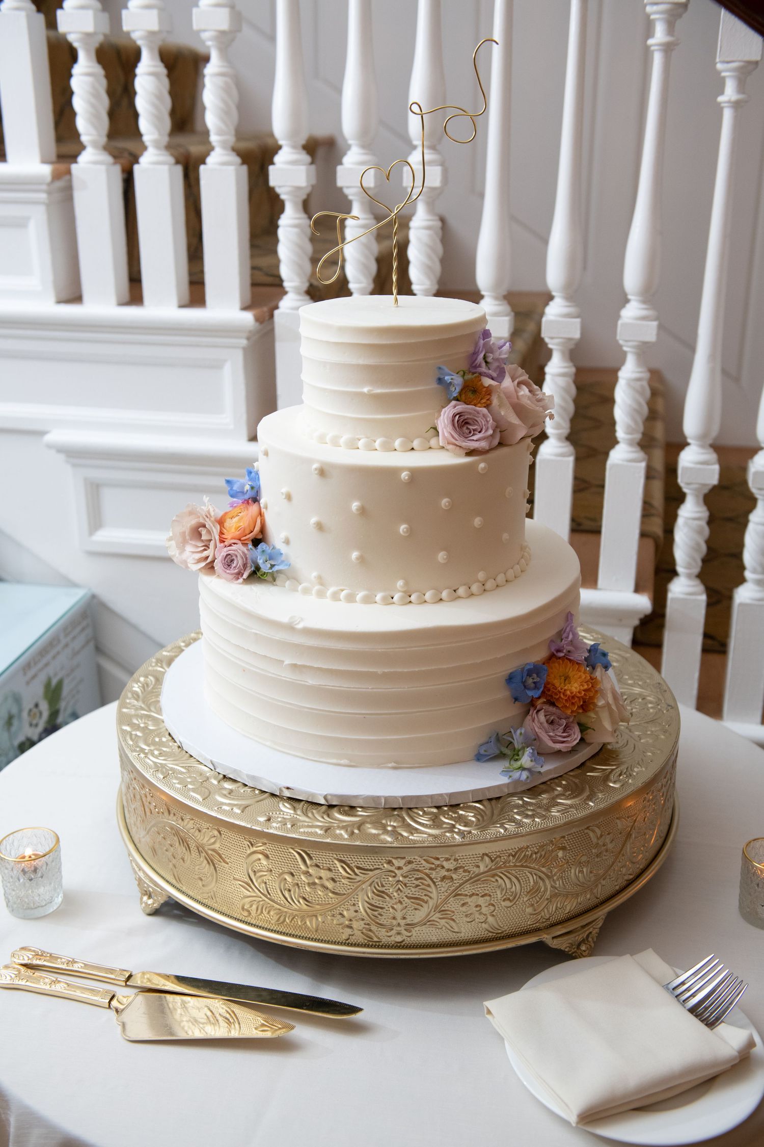 White wedding cake with delicate floral decorations sits on an ornate gold cake stand against white railing backdrop.