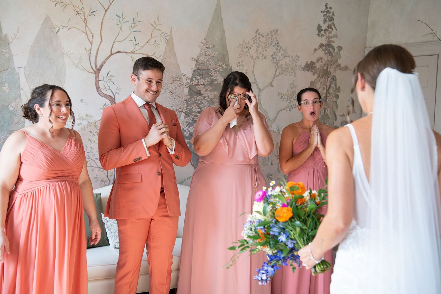 Wedding party members in coral and peach colored dresses gather for a joyful group photo in an elegant indoor setting.