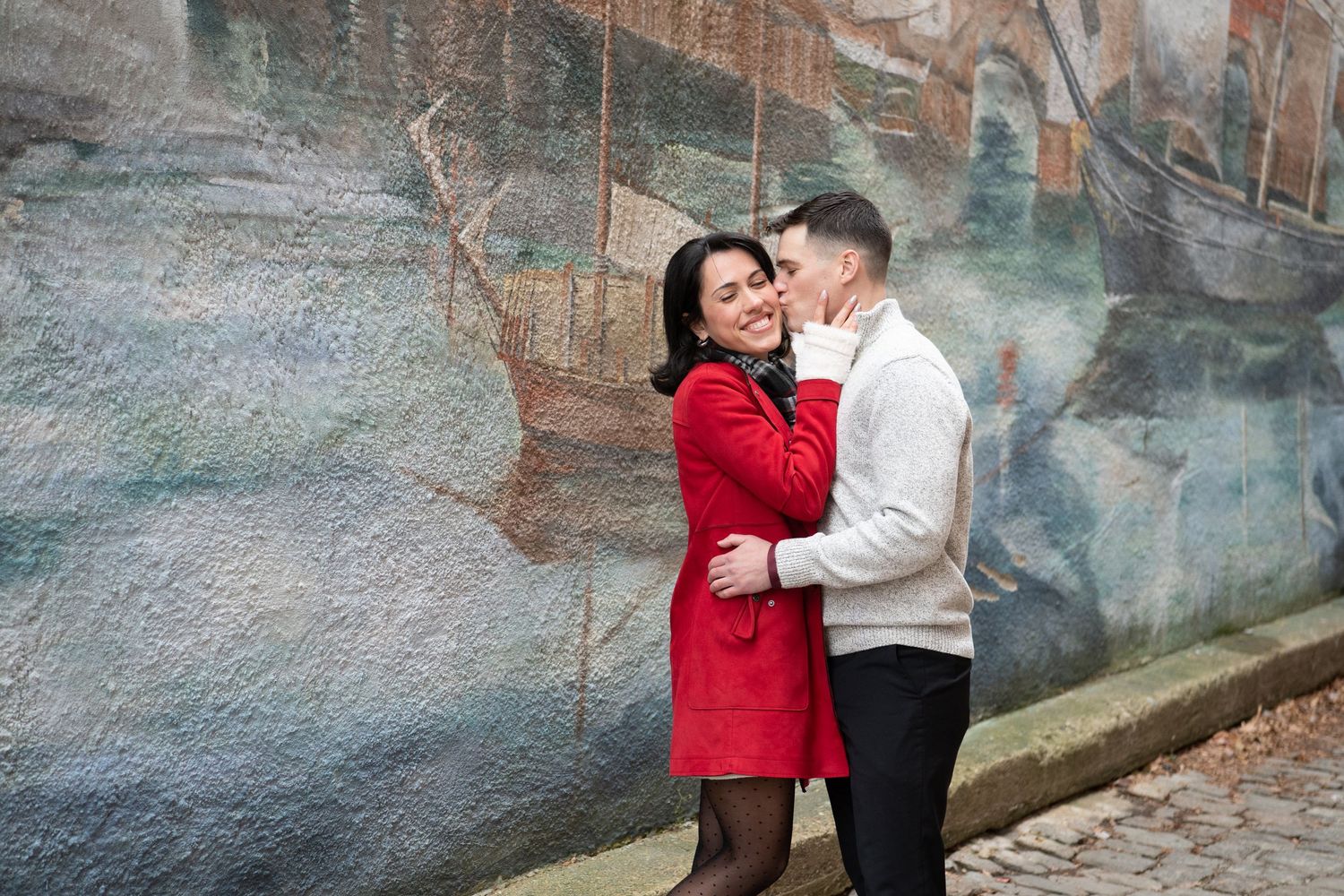 A couple in winter attire shares romantic moments against a rustic stone wall backdrop during a photo session.