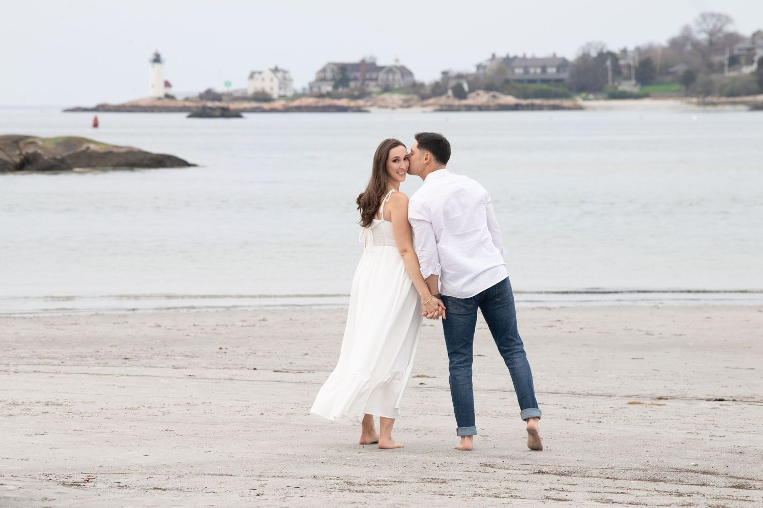 A romantic coastal scene with a couple embracing on a sandy beach with a lighthouse in the distance.