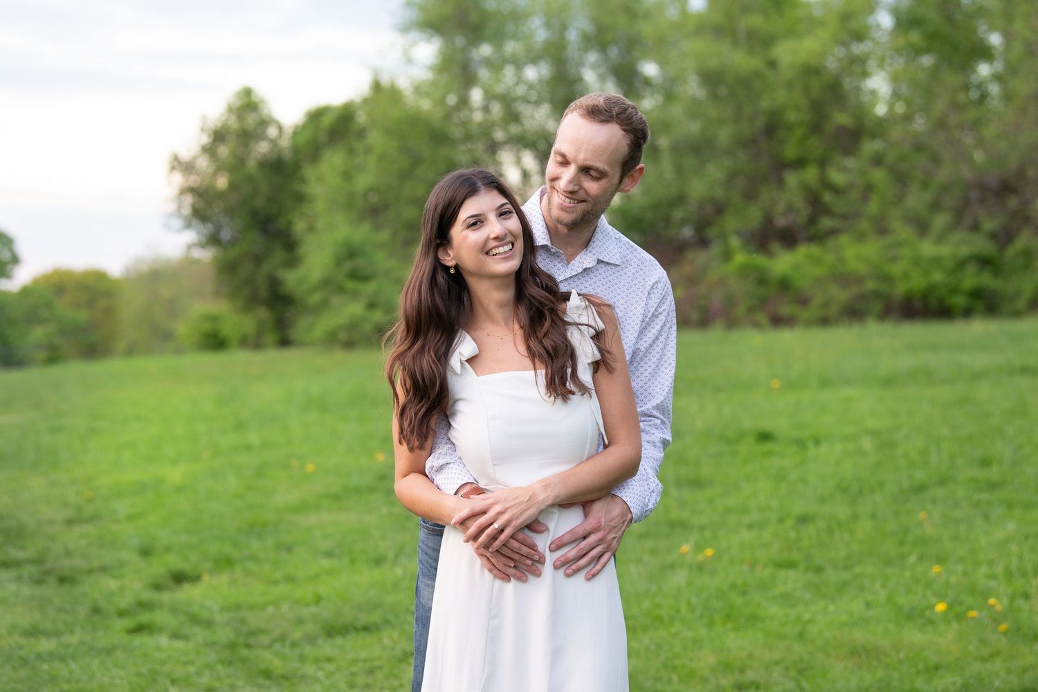 A couple embraces on a lush green hillside while wearing coordinated white outfits during a sunny afternoon.