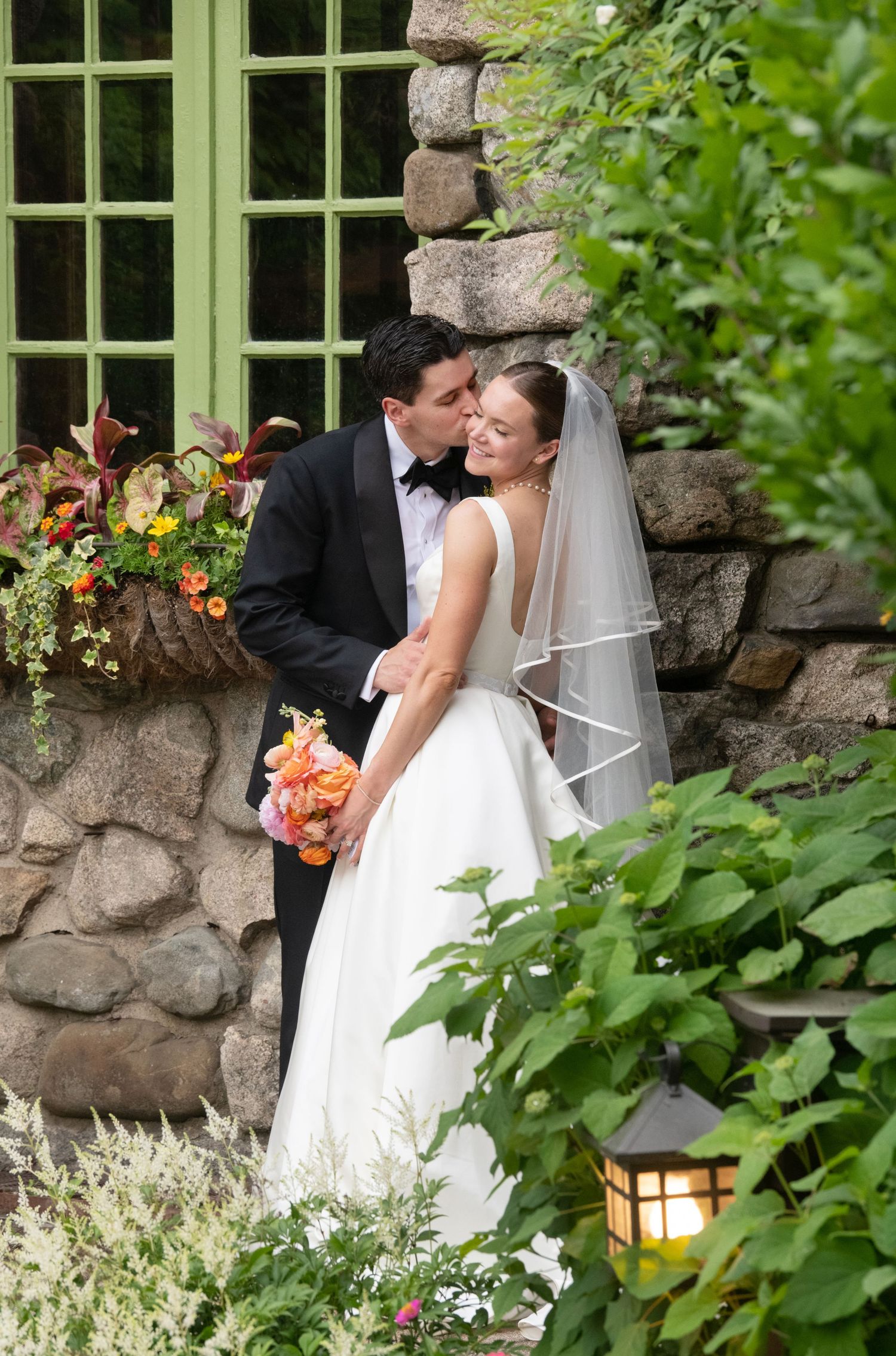 Groom in tuxedo kissing bride in white gown and veil, holding orange bouquet, beside stone wall with floral window box.
