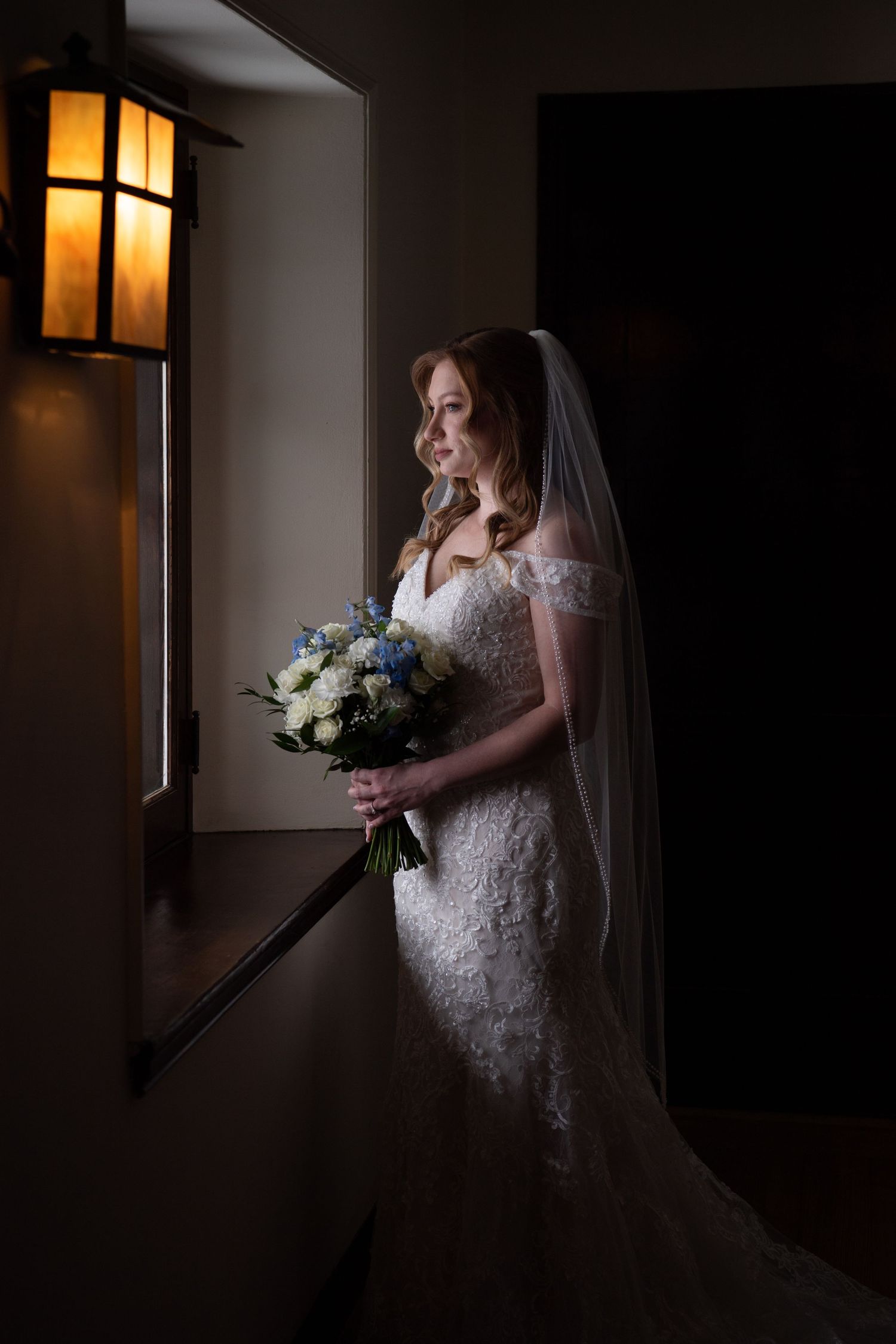 A bride in a lace wedding dress poses in dramatic window light holding a white and blue floral bouquet.