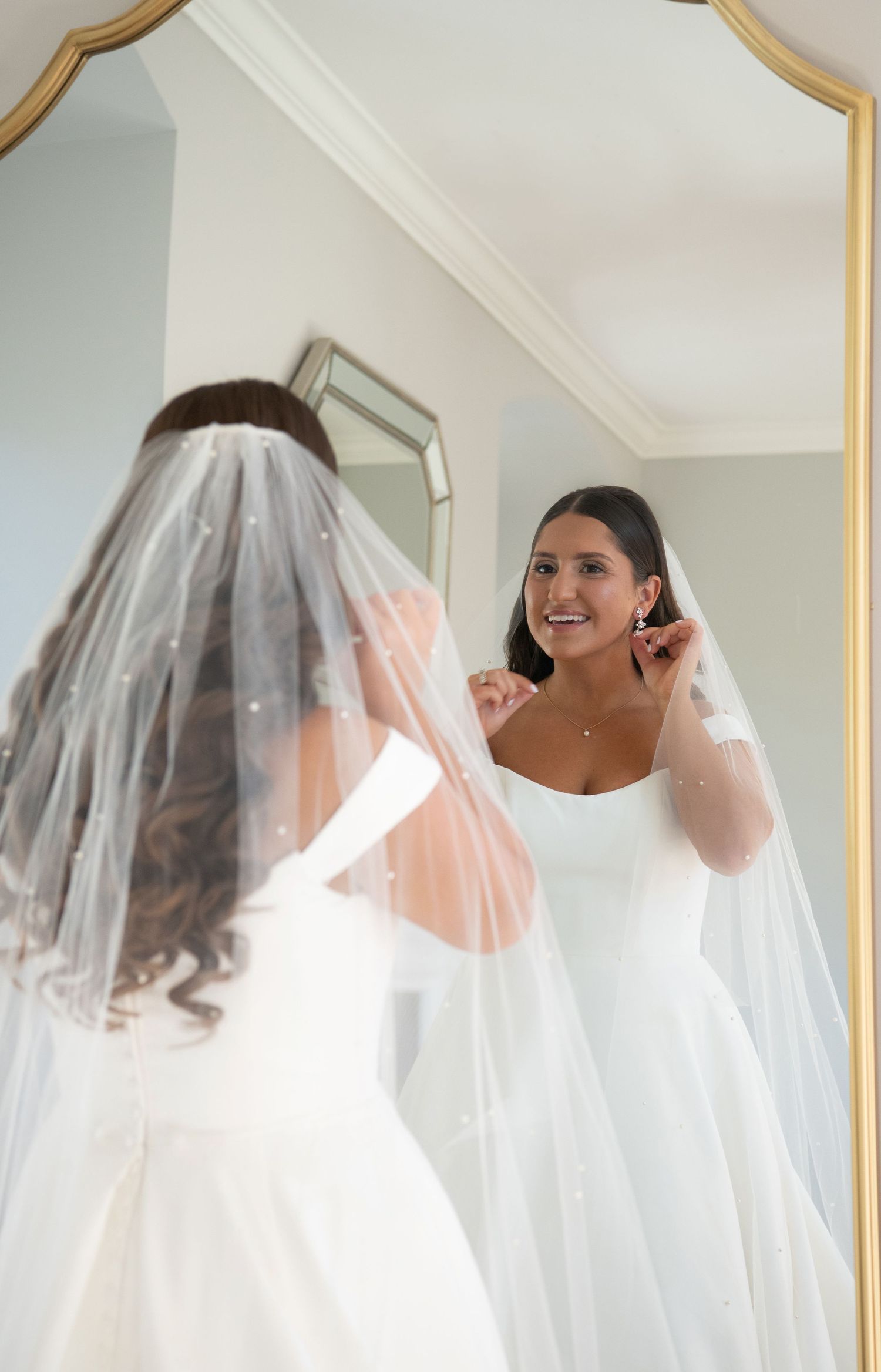 A bride in a white wedding dress and veil smiles while looking at her reflection in an ornate gold-framed mirror.