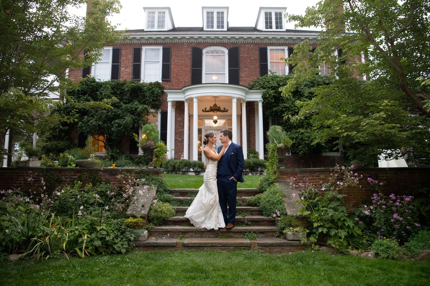 Wedding party poses in front of a grand colonial-style mansion with landscaped gardens.
