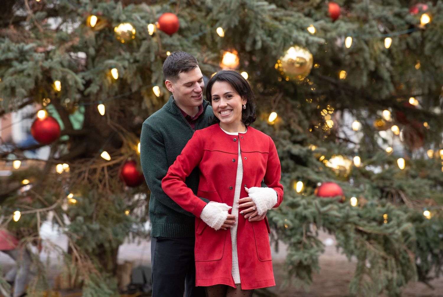 A couple embraces in front of a decorated Christmas tree with red and gold ornaments and twinkling lights.