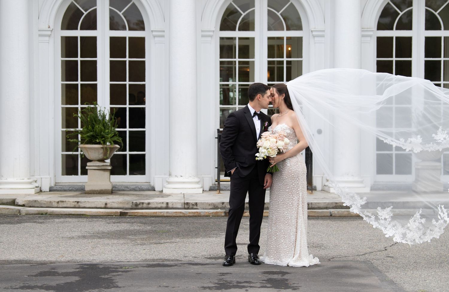 Bride and groom share a romantic kiss outside elegant white venue, veil flowing in the wind, bouquet in hand.
