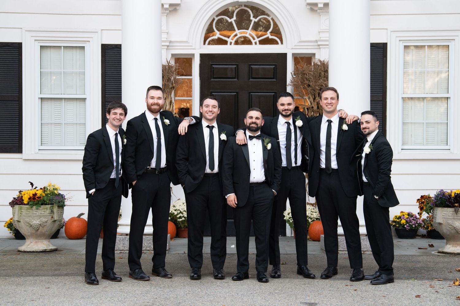 A group of groomsmen in black tuxedos and bow ties stand together in front of a white building with an arched doorway.