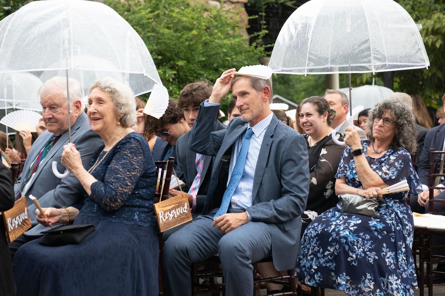 Wedding guests hold clear umbrellas during outdoor ceremony.