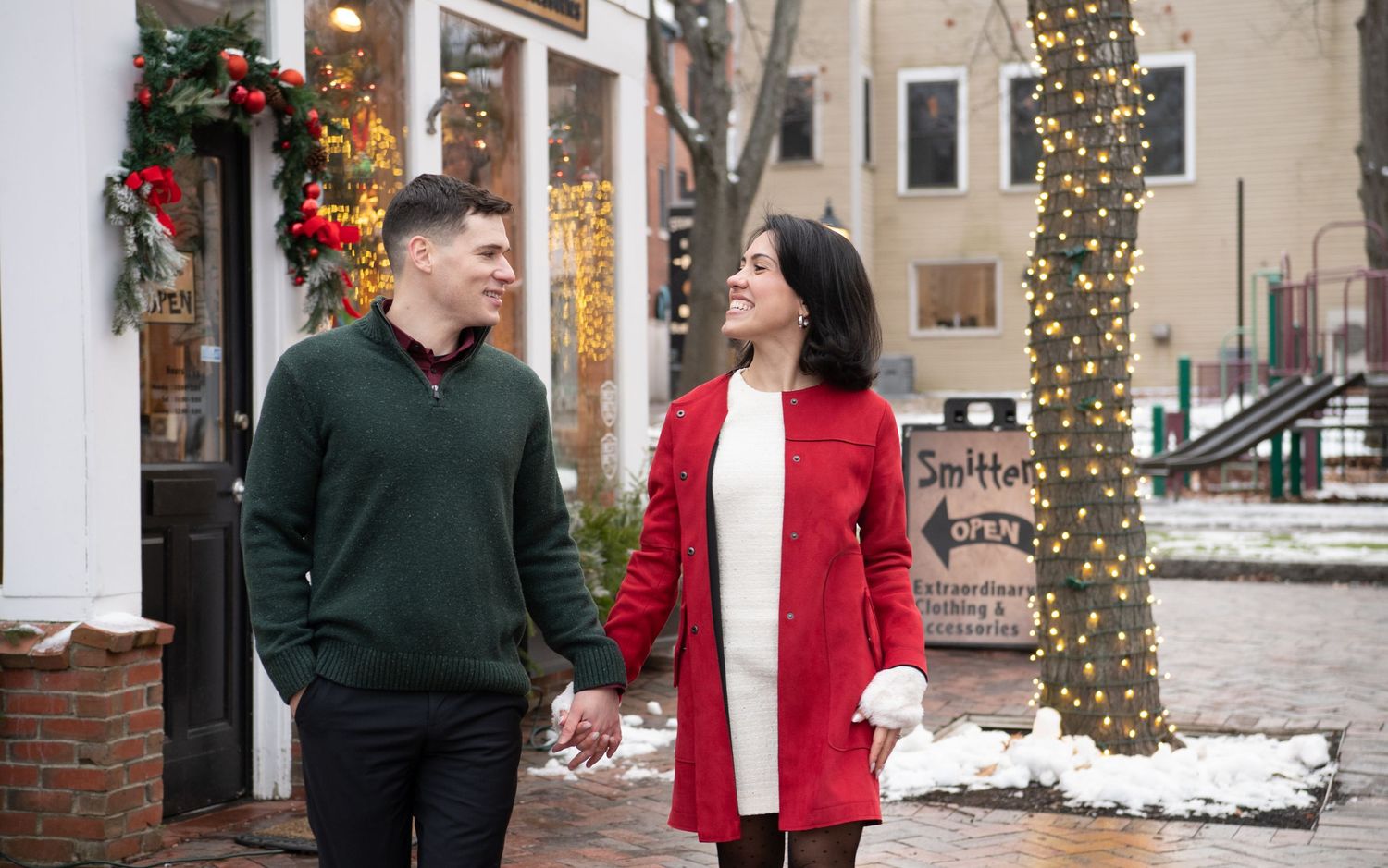 A couple in winter coats share a romantic moment outdoors with snow and holiday decorations in the background.