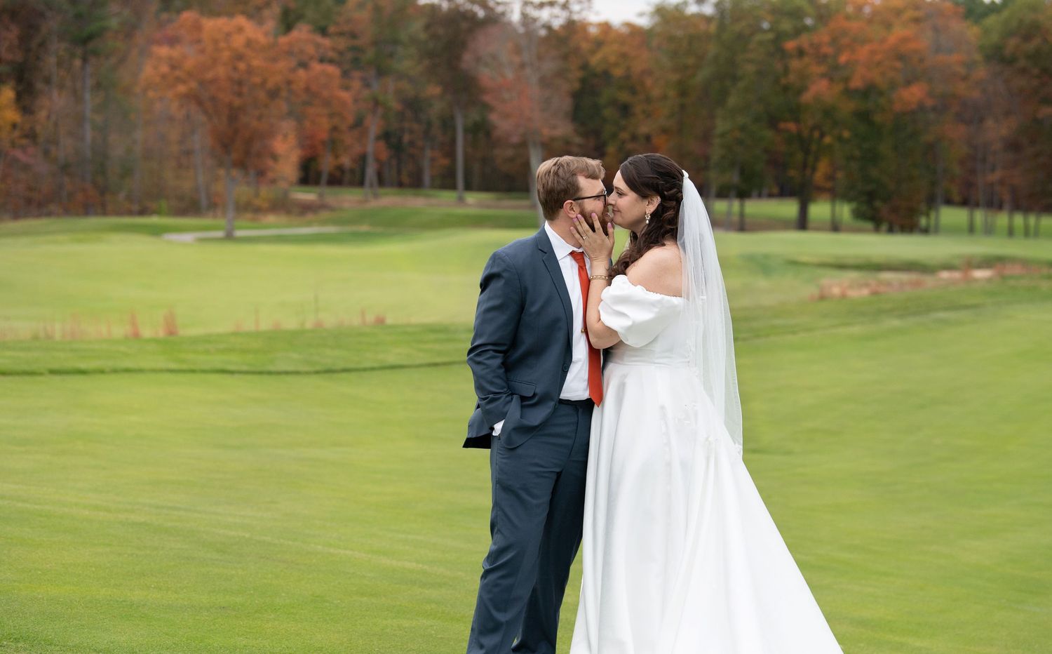A couple shares a romantic moment on a golf course with autumn trees in the background during their wedding photoshoot.