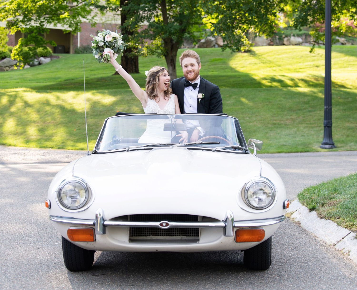 A classic white Jaguar E-Type convertible with newlyweds drives down a tree-lined path on a sunny day.