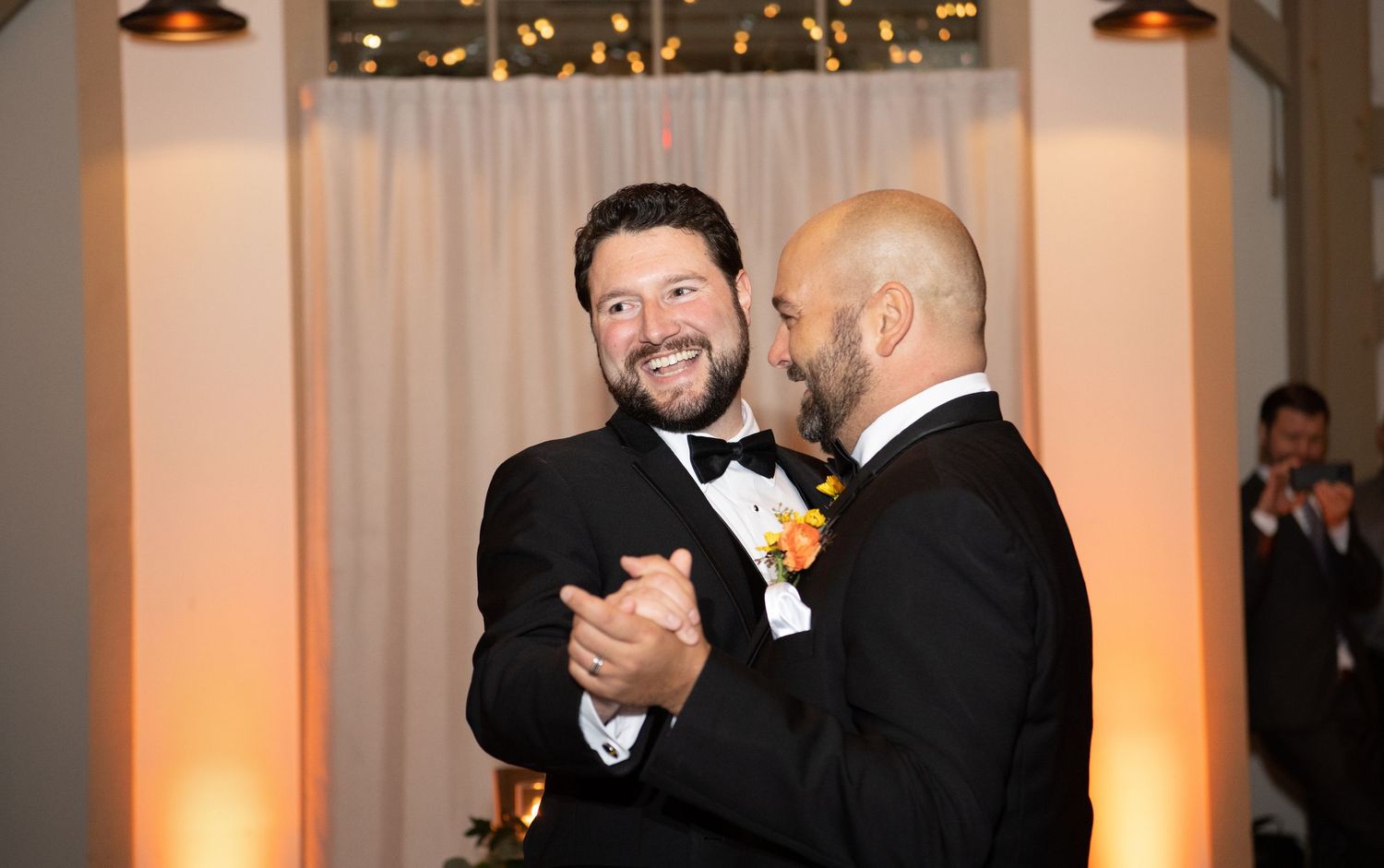 Two people embrace in a joyful moment at an indoor wedding reception with warm lighting and curtains in the background.