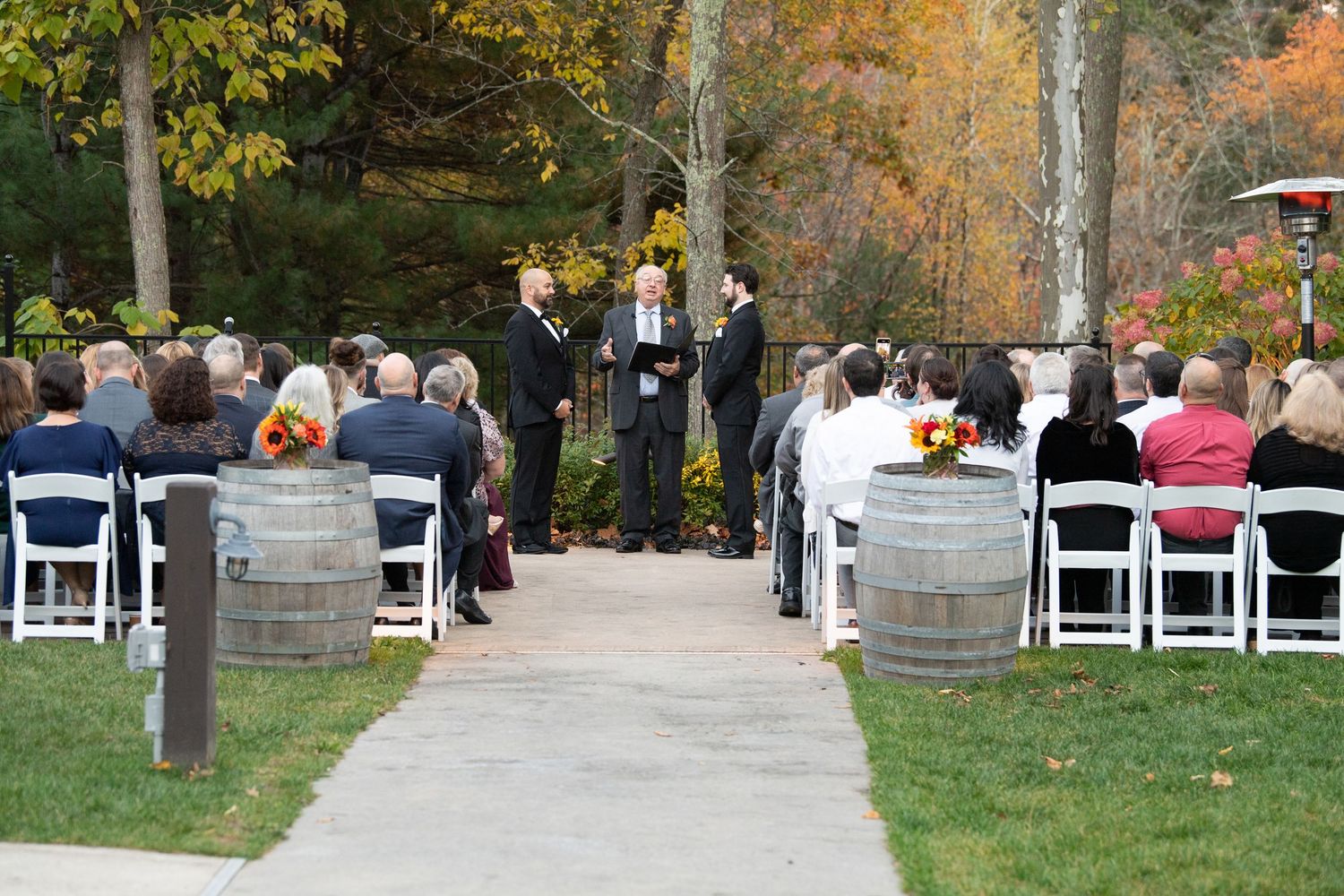 Guests seated in white chairs at an outdoor autumn wedding ceremony with colorful fall foliage in the background.