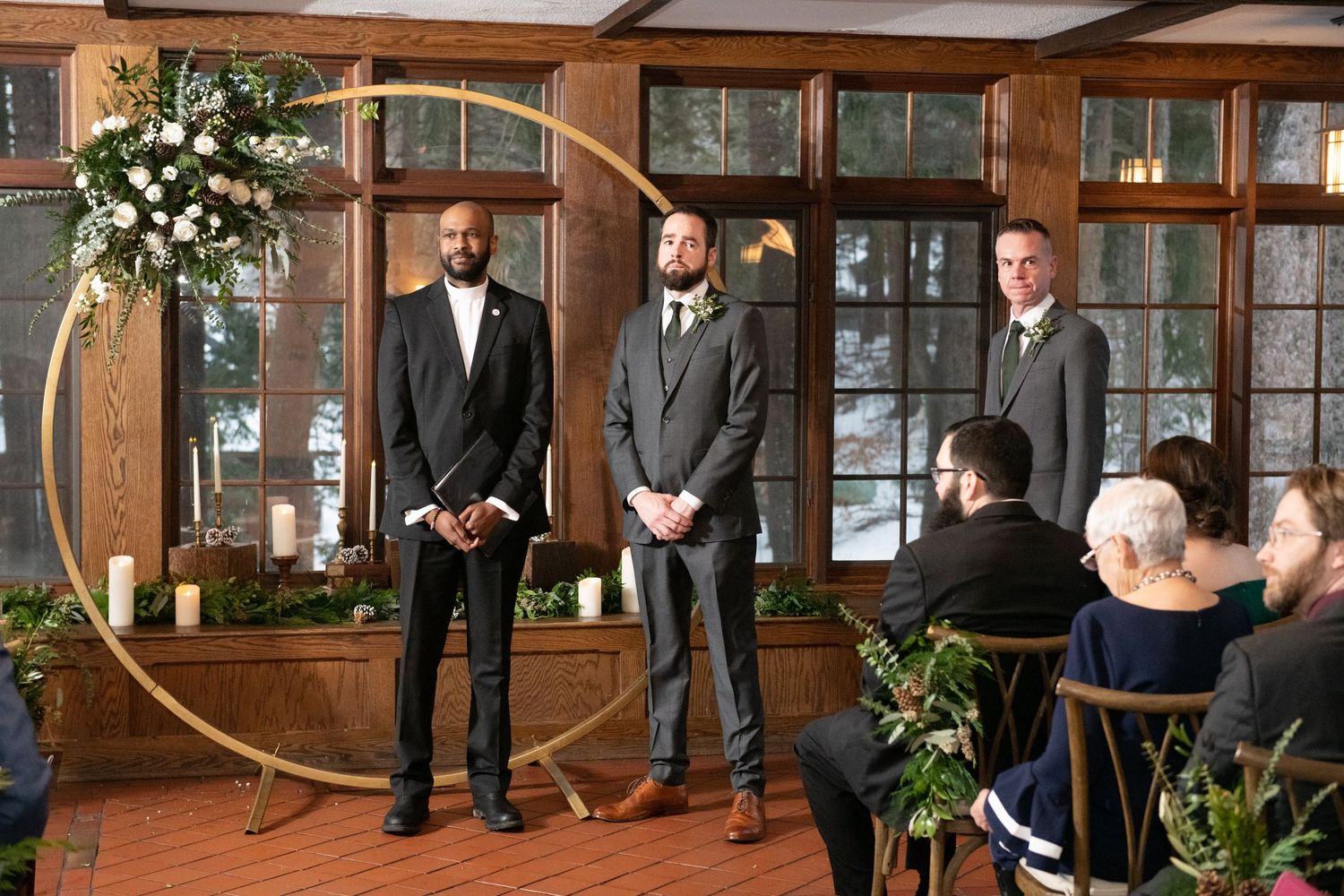 Three men in formal suits stand at the front of an indoor ceremony space with wooden beams and floral decor.