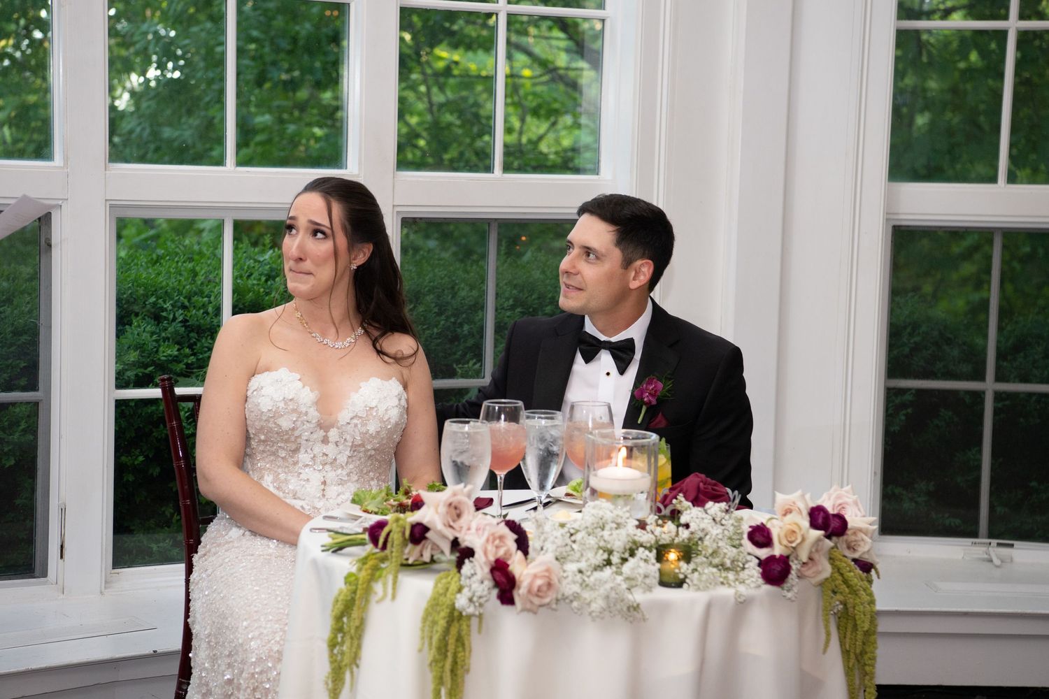 Bride and groom seated at sweetheart table with floral centerpiece during wedding reception.