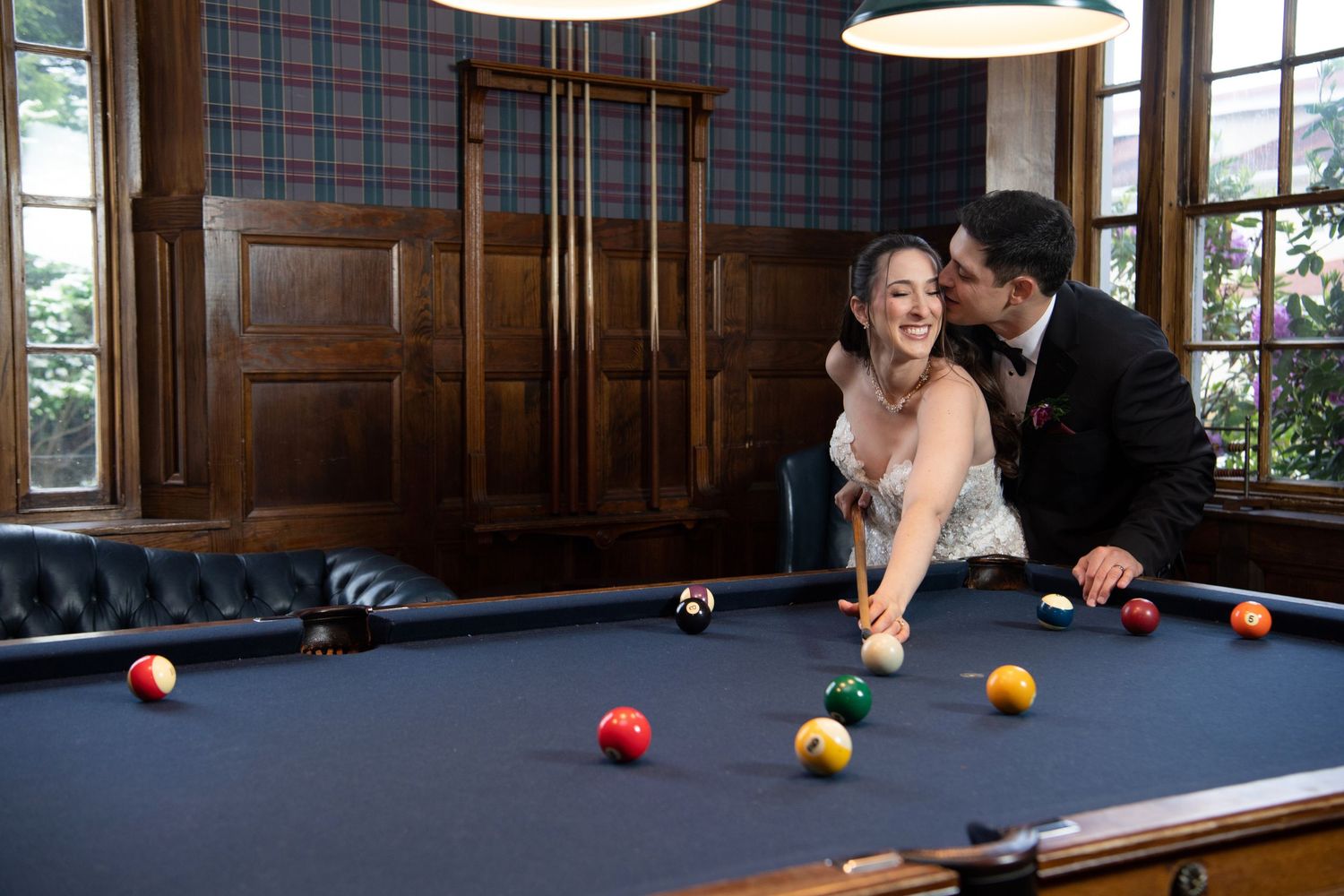 Bride and groom share a kiss while playing pool on their wedding day in an elegant wood-paneled billiards room.