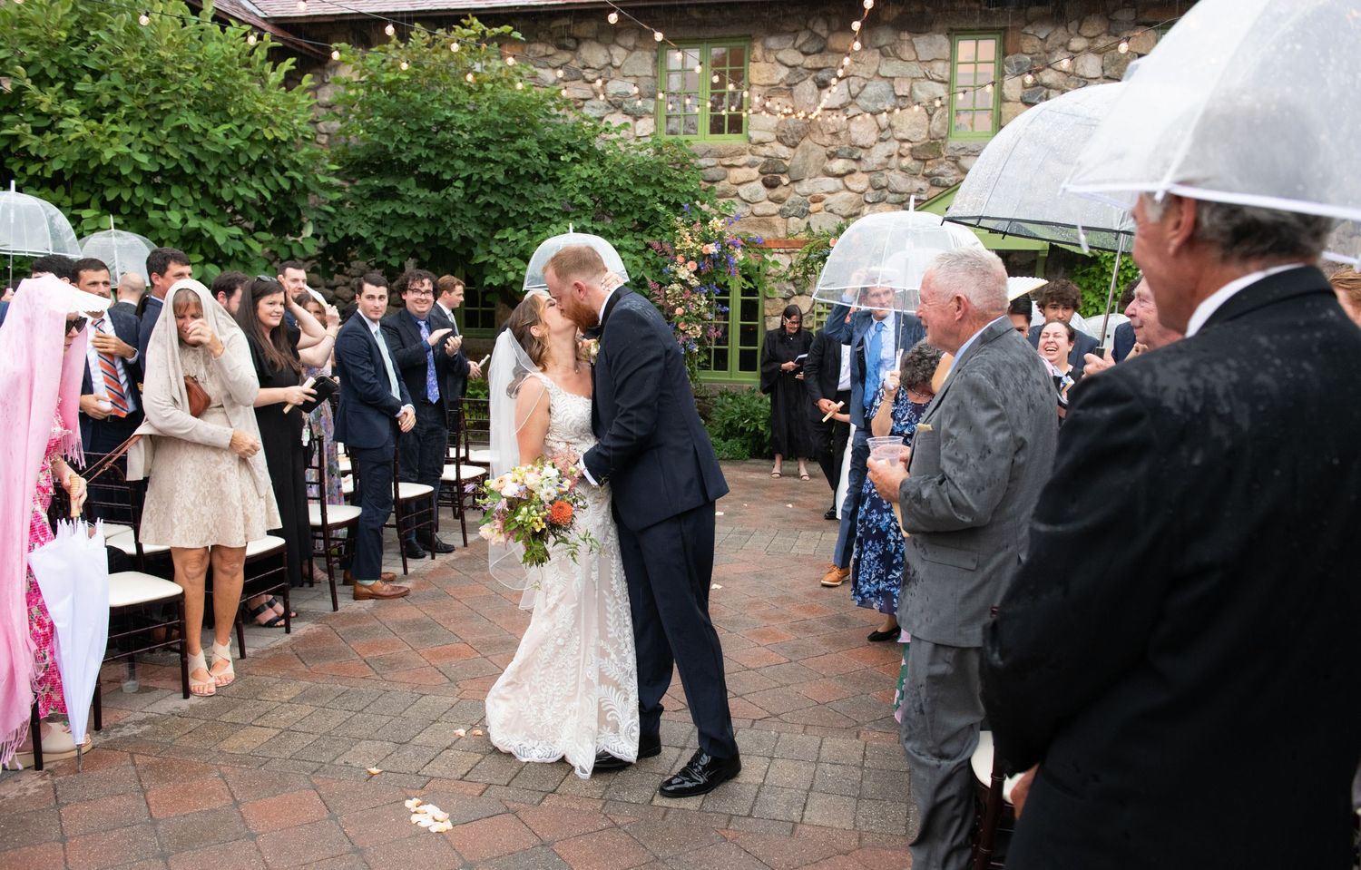 A couple shares their first kiss at an outdoor wedding ceremony while guests look on holding umbrellas in the rain.
