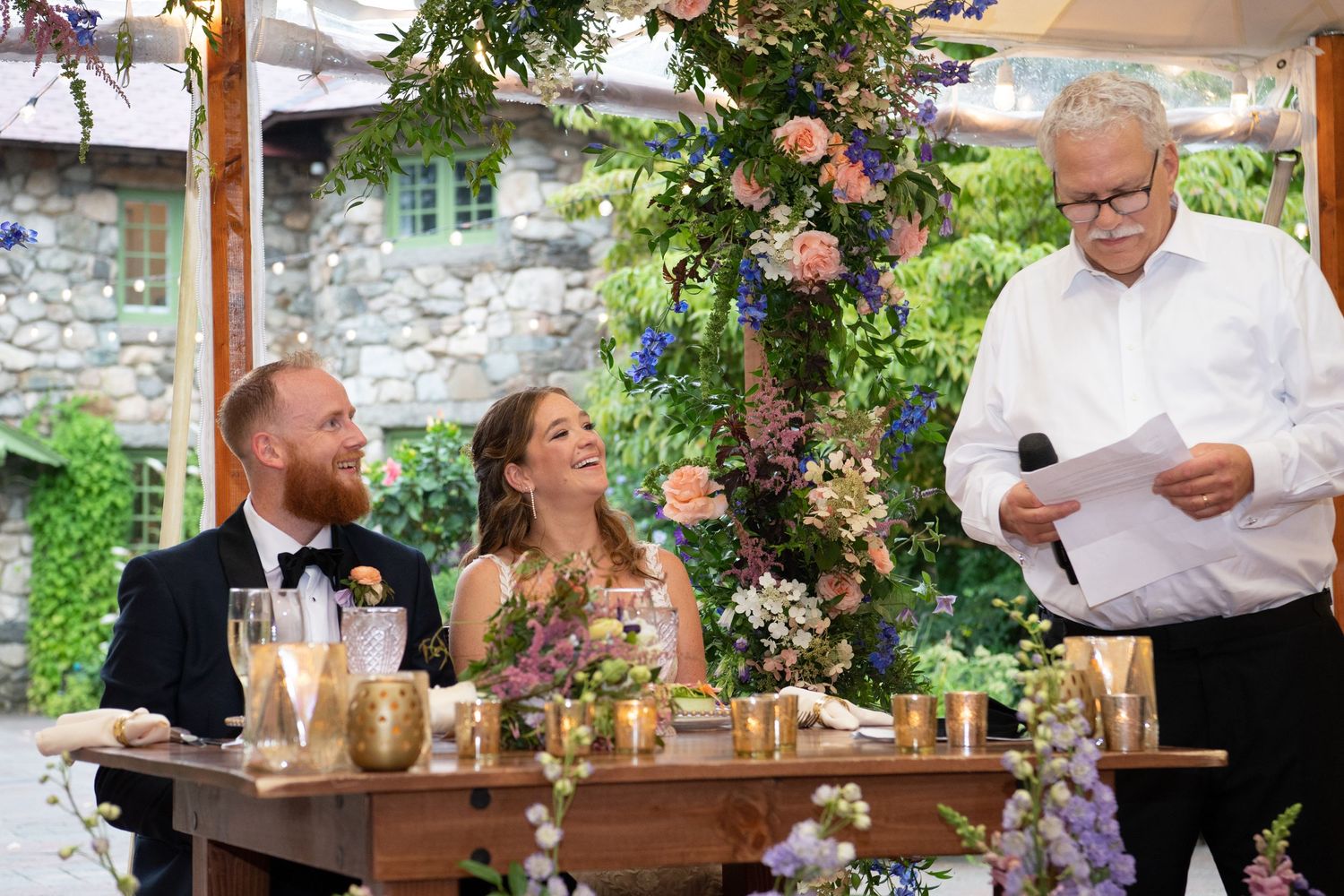 Wedding guests seated at rustic wooden tables decorated with floral arrangements under outdoor tent.