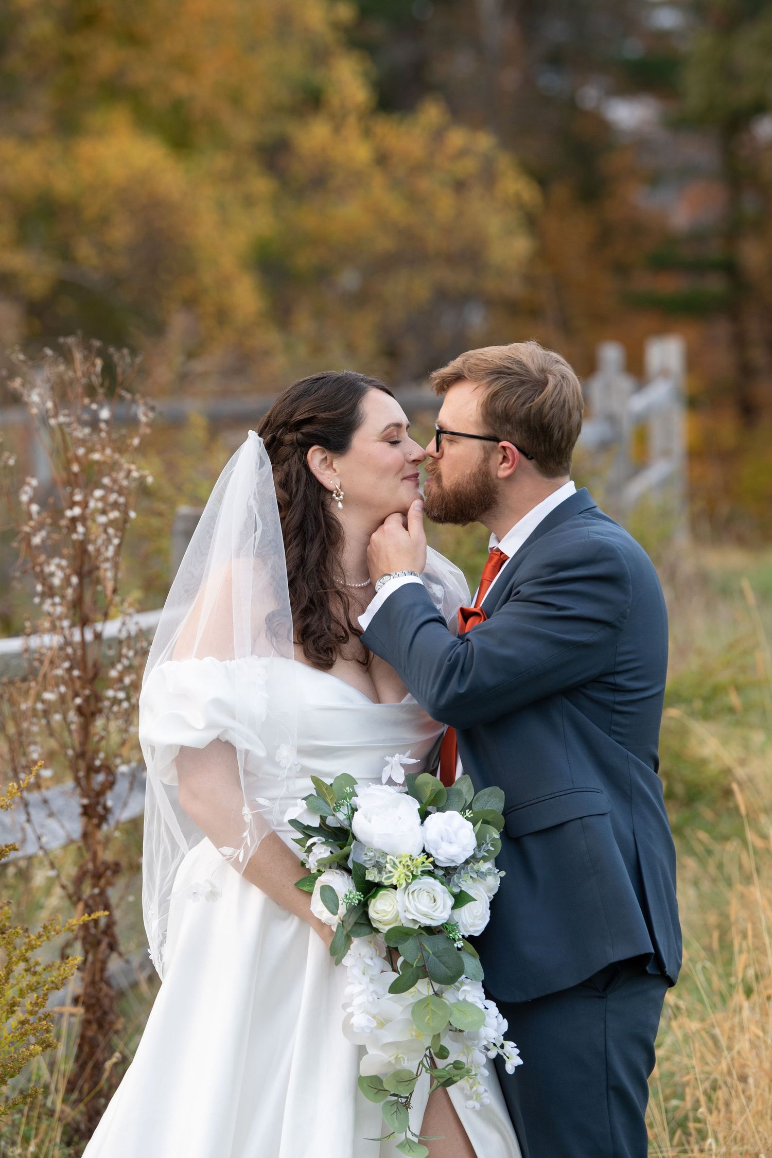 Newlywed couple shares romantic kisses in a fall meadow with white bridal bouquet and flowing veil.