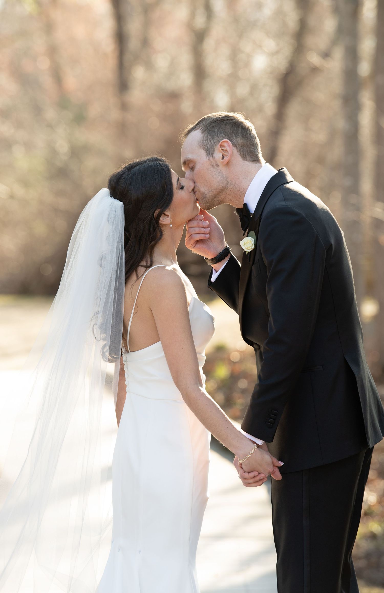 Bride and groom sharing a romantic kiss while holding hands outdoors, bride wearing white gown and veil.