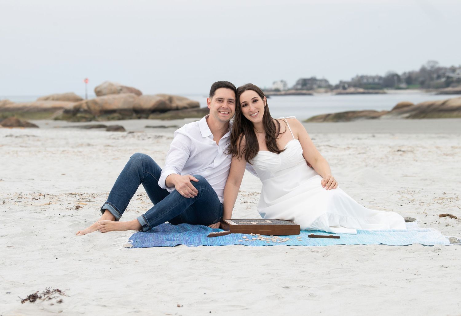 A couple in casual and formal attire sit together on a blue blanket at a rocky beach on an overcast day.