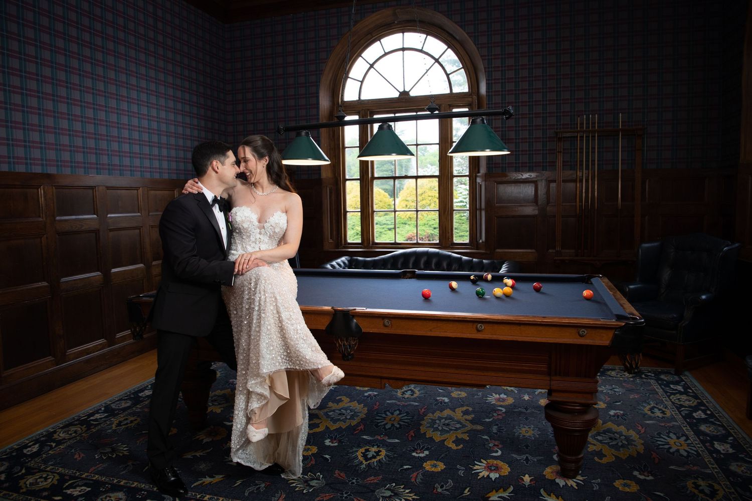 Bride and groom share a romantic moment in an elegant billiard room with wood paneling and arched windows.