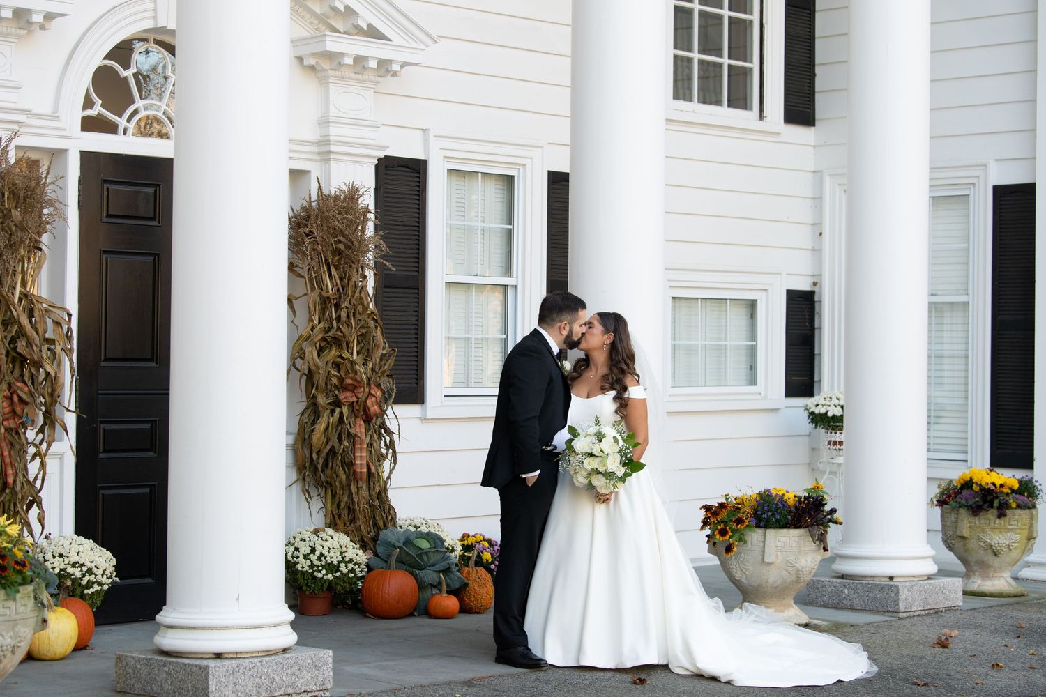 A white colonial-style porch decorated with fall pumpkins, corn stalks and wreaths creates an elegant autumn entrance.