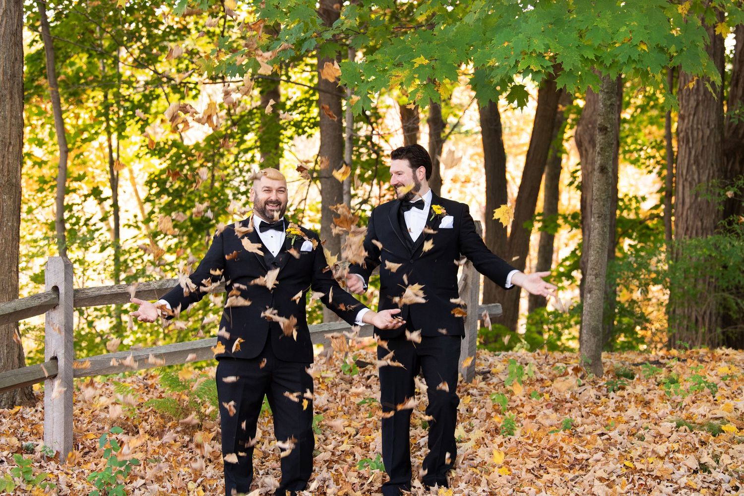 Wedding party in black formal attire poses together among autumn trees with golden leaves in an outdoor setting.