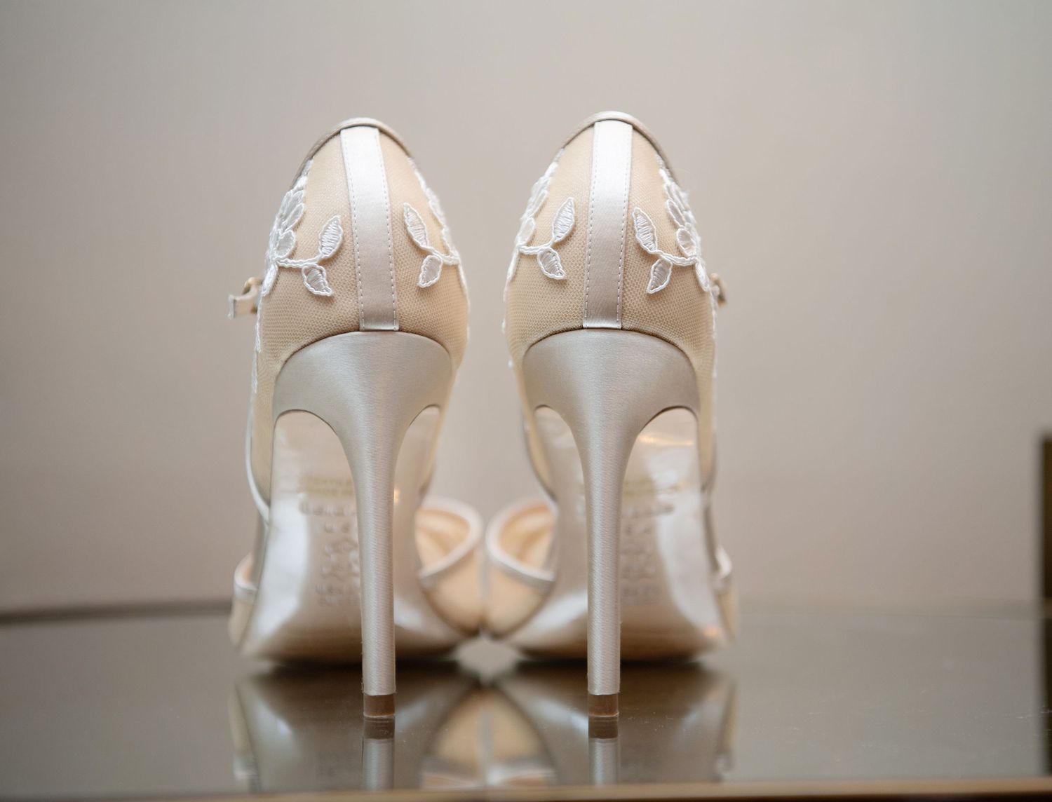 Elegant white bridal high heels with delicate floral embellishments shown from behind on reflective surface.