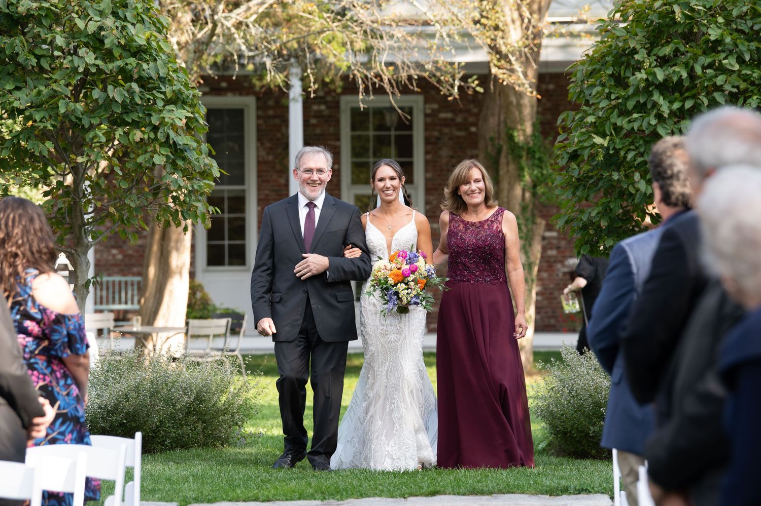 Wedding party walking outdoors through a garden setting with lush greenery and trees.