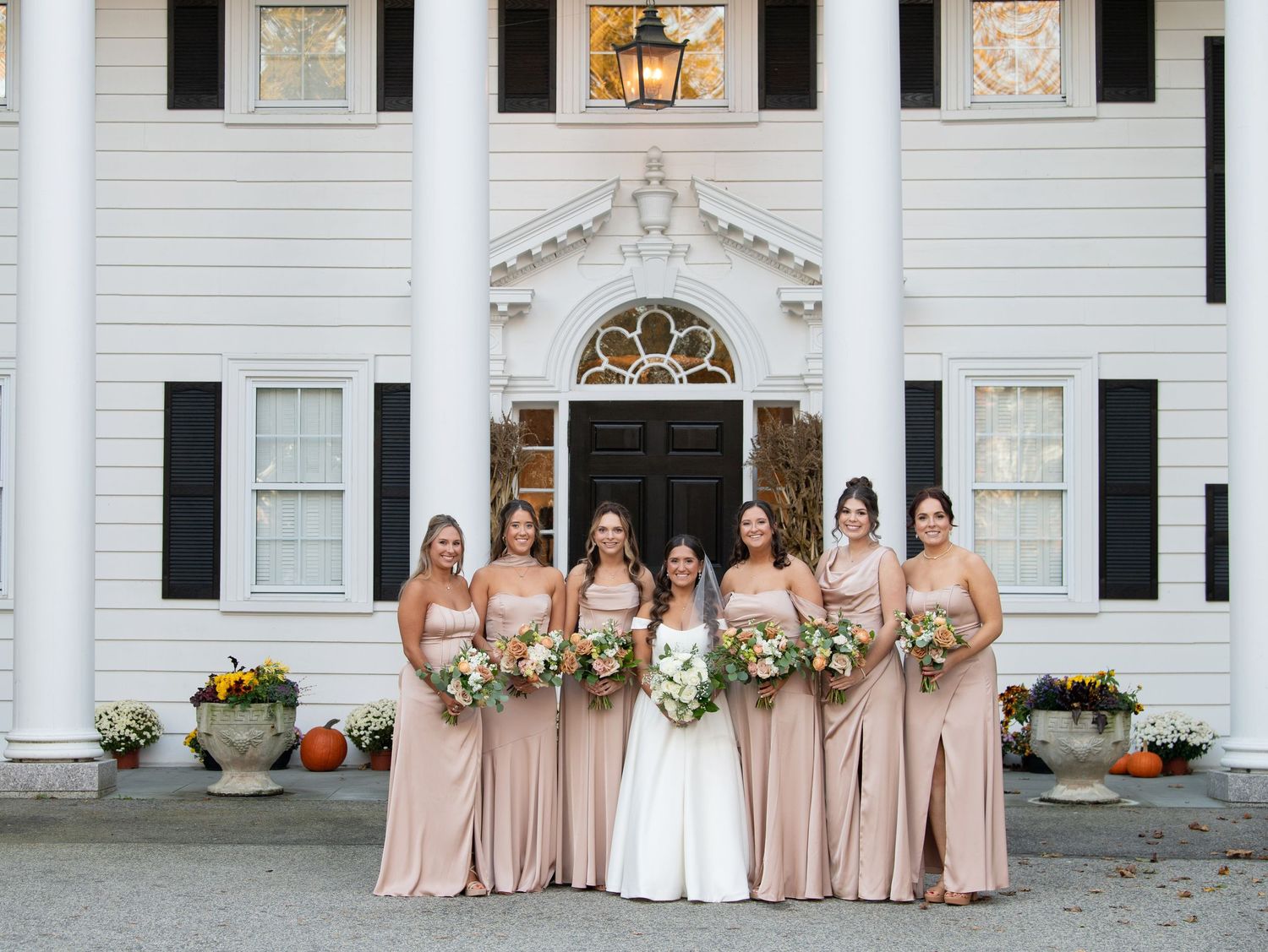A wedding party poses in front of a grand white mansion with elegant columns and decorative architectural details.