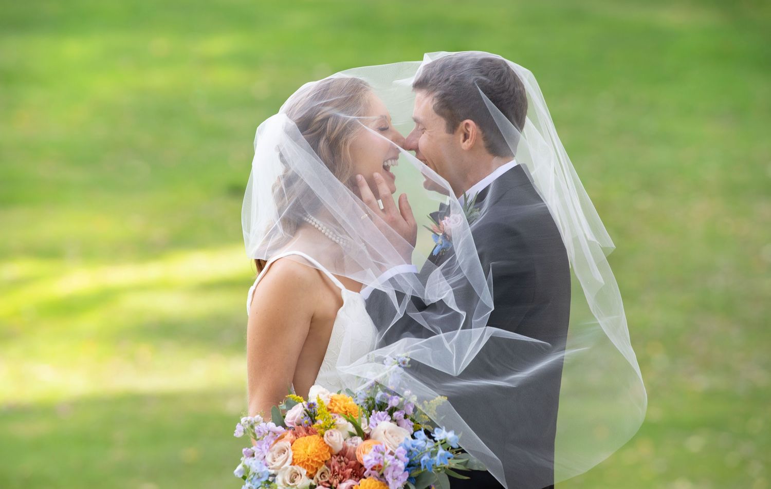 A romantic wedding couple embraces under a veil in a lush garden setting with soft natural lighting.