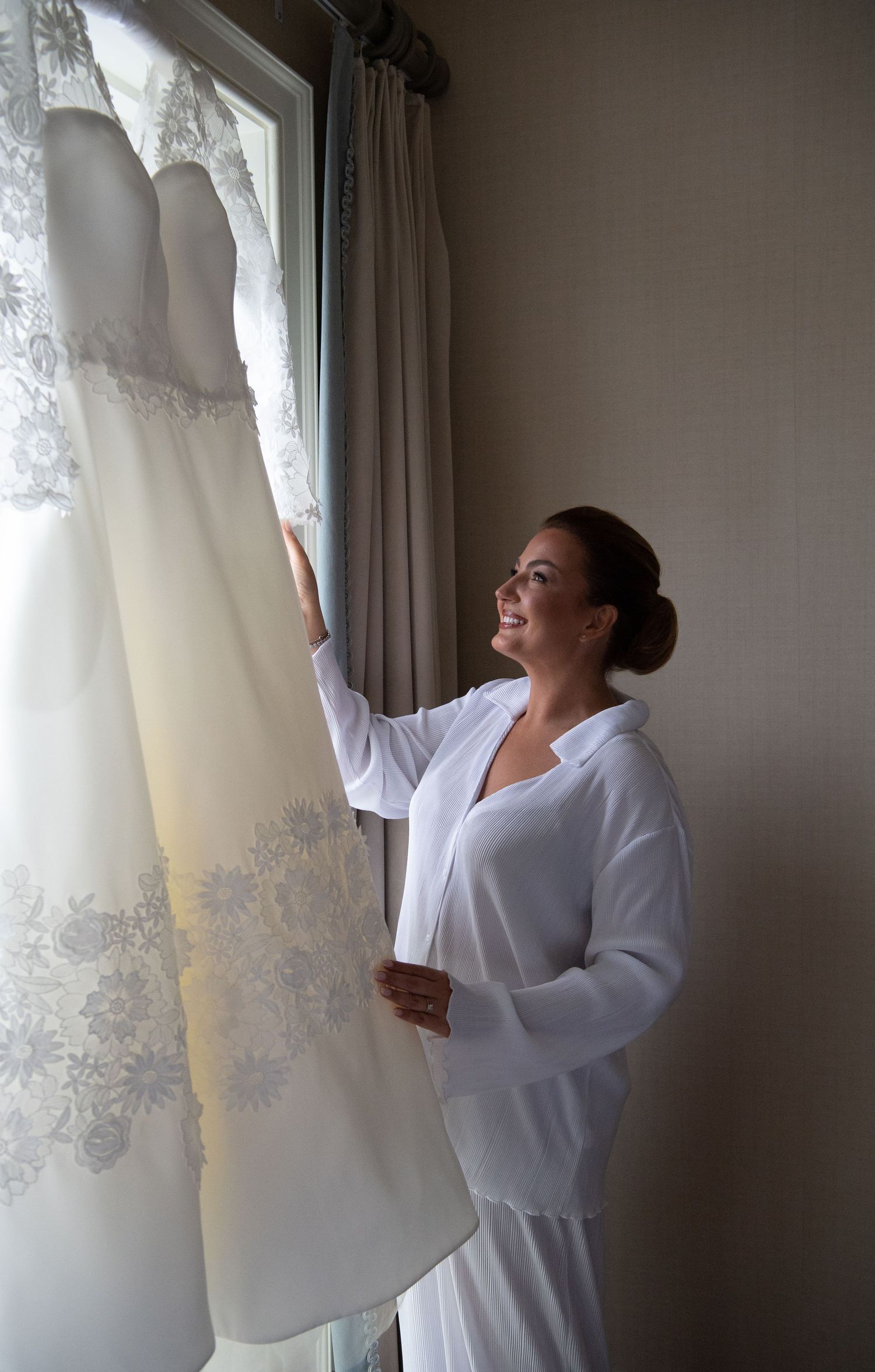 A woman in a white robe examines a delicate lace wedding dress hanging by a window in soft natural light.