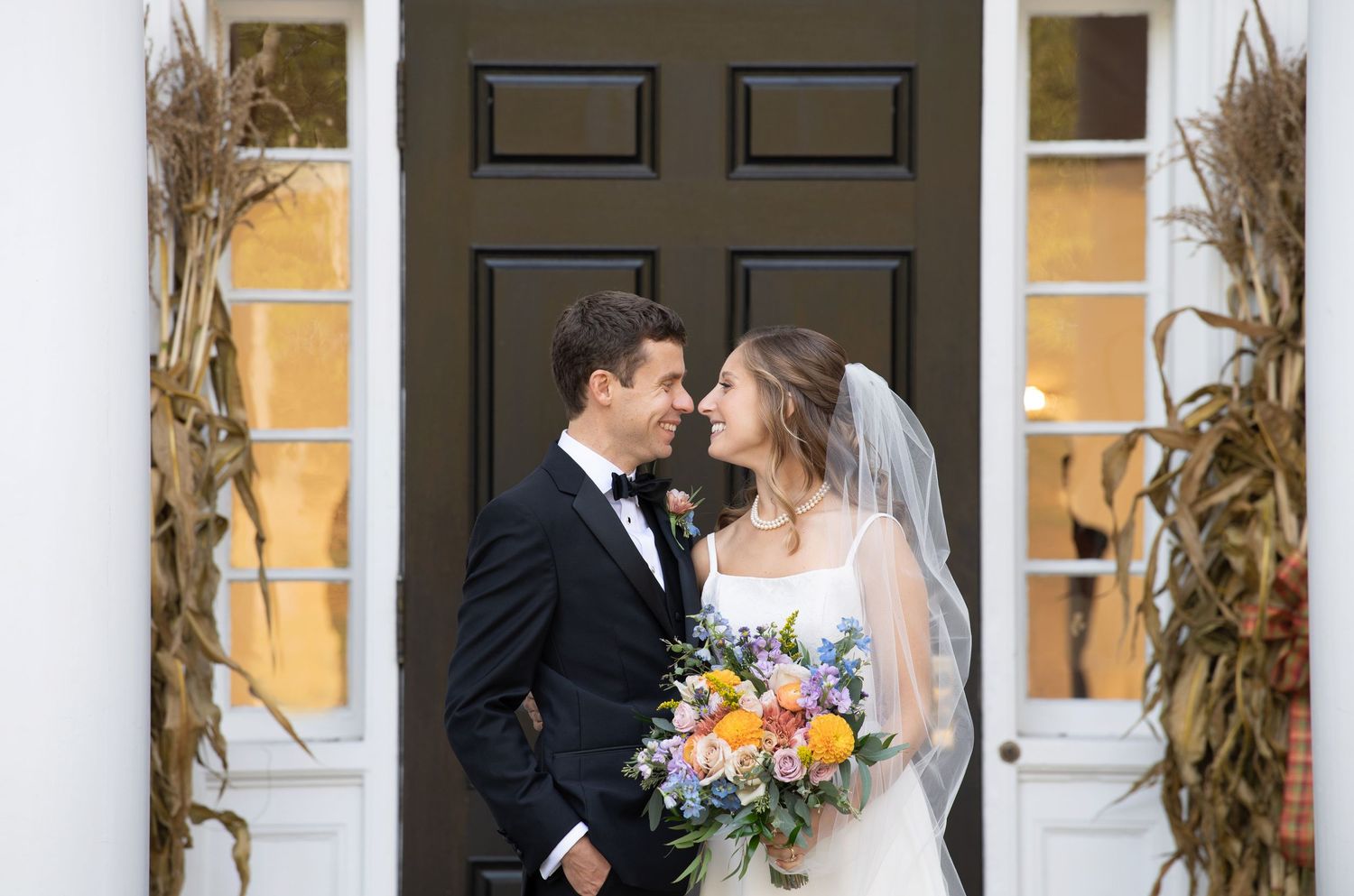 A couple in wedding attire share romantic moments in front of a yellow building with decorative cornhusks and windows.