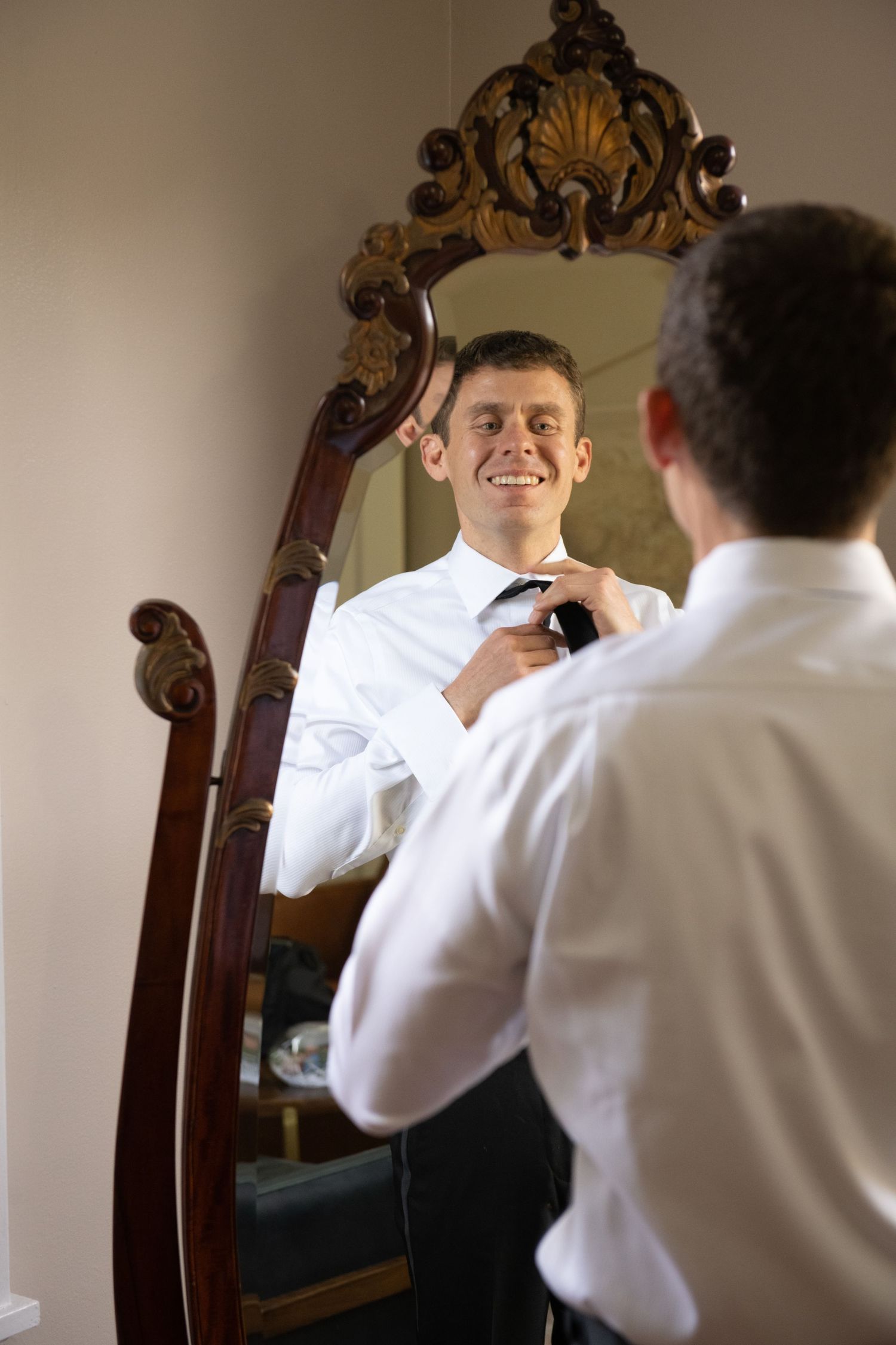 A person adjusts their tie while looking in an ornate mirror during wedding preparations.