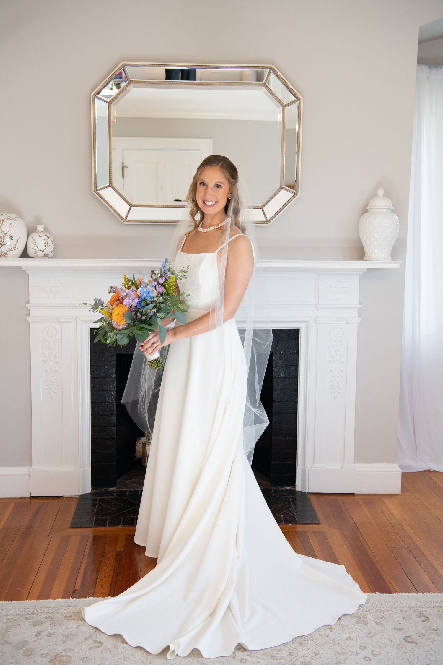 A bride in a flowing white wedding gown twirls and poses in a bright room with hardwood floors and a fireplace.