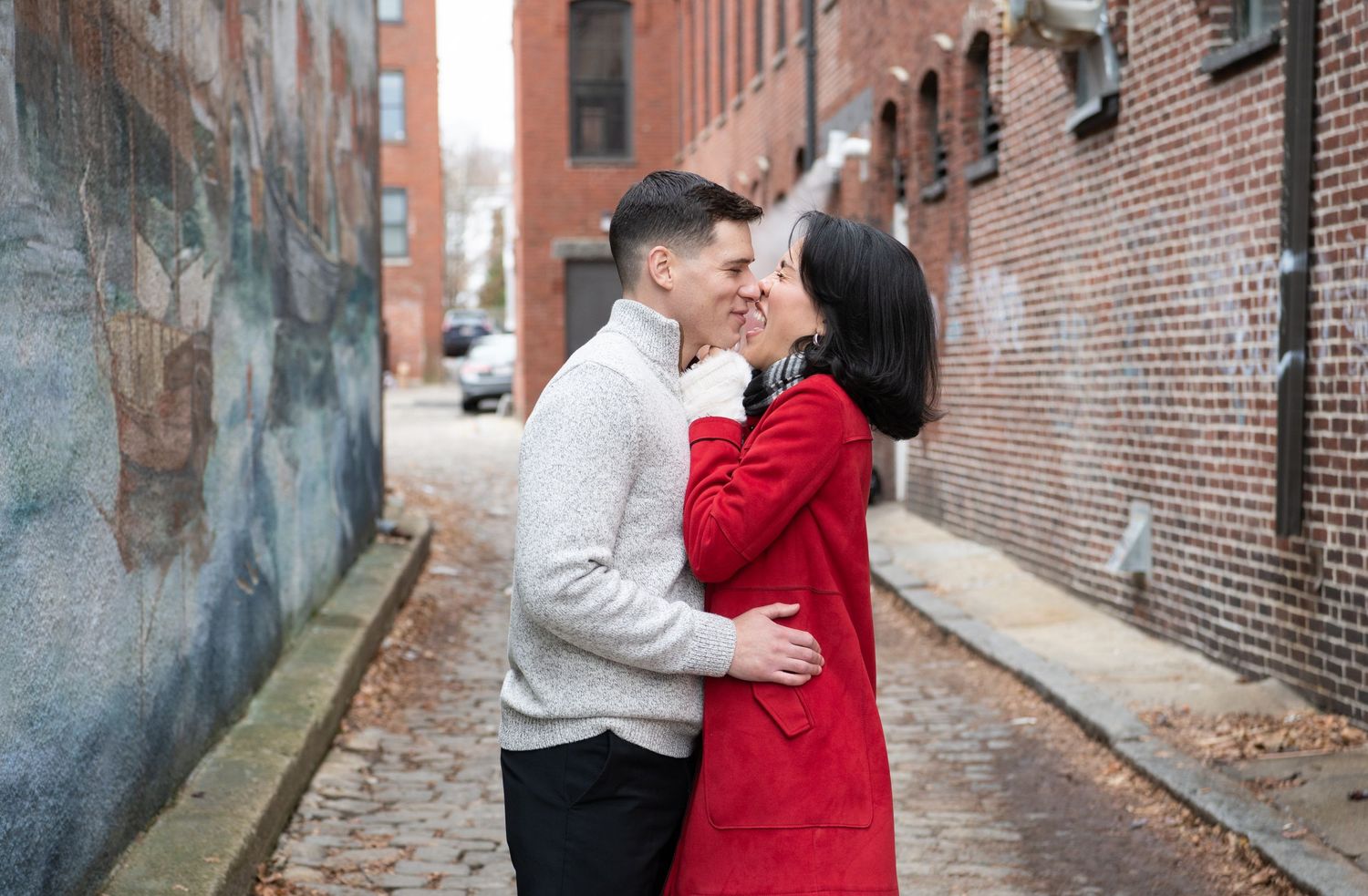 A couple shares a romantic moment on a cobblestone street against a brick wall, with one wearing a red coat and white sweater.