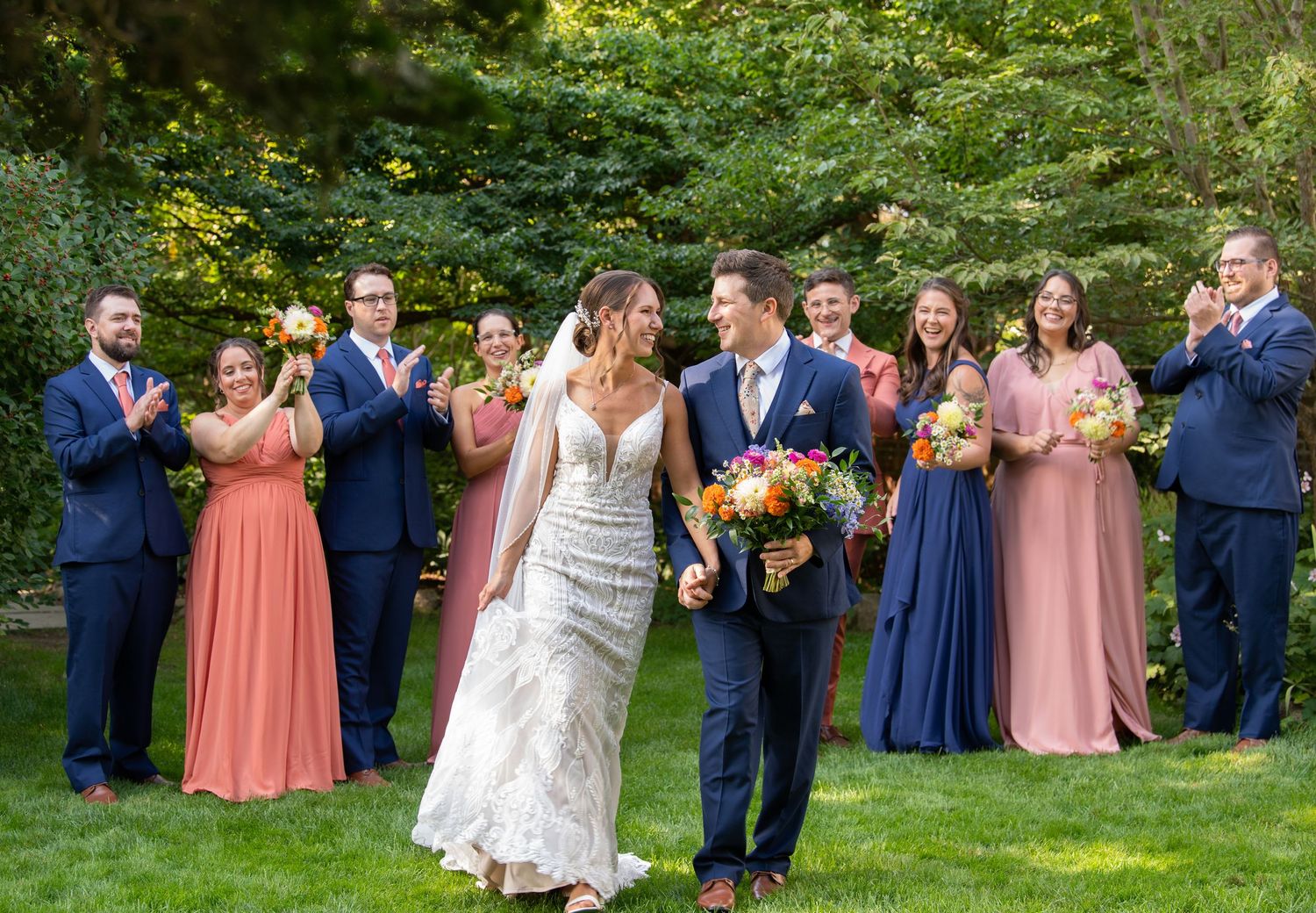 Wedding party celebrates outdoors with bridesmaids in coral dresses and groomsmen in navy suits during summer ceremony.