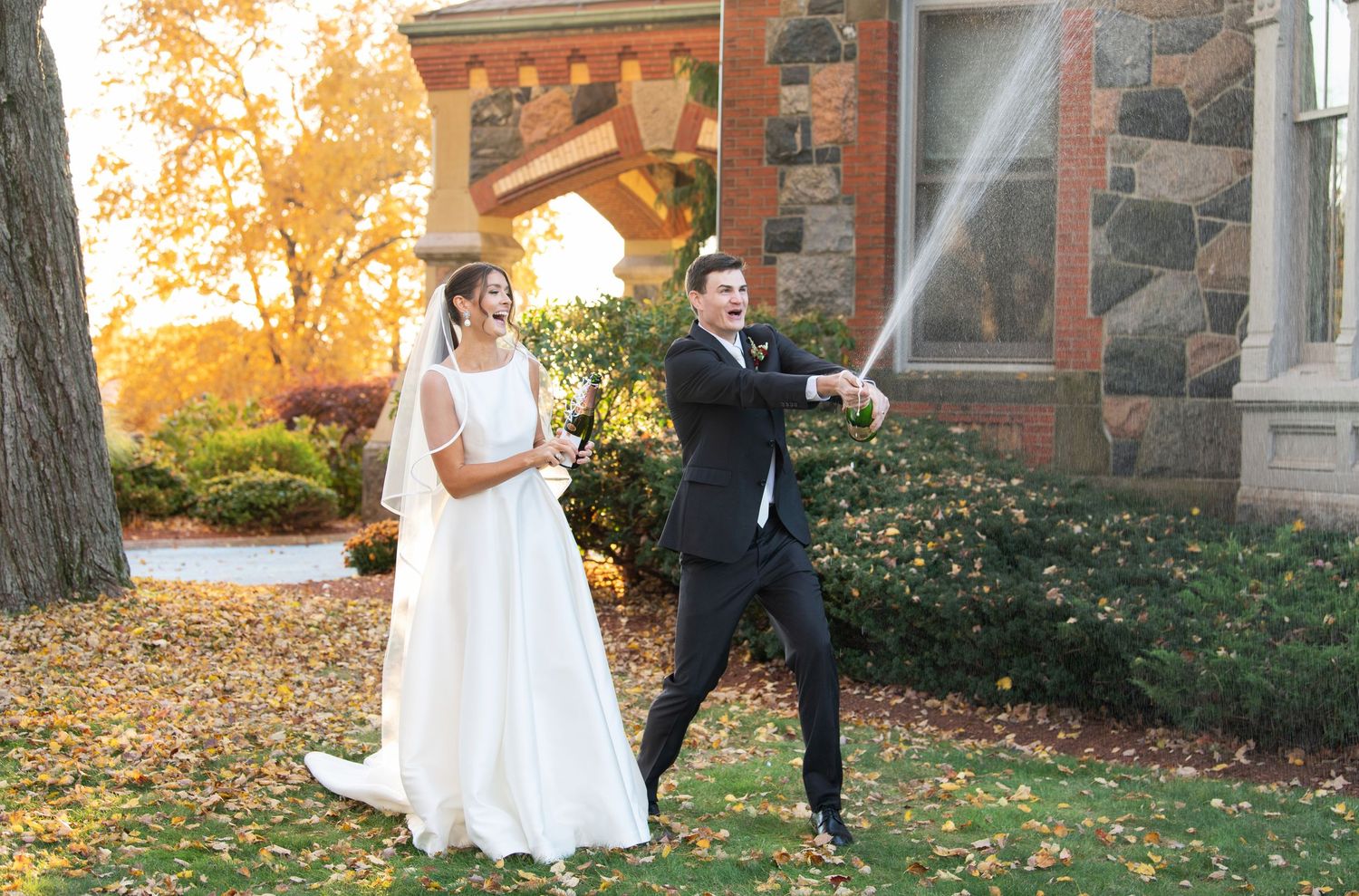 A bride and groom walk together on a stone path surrounded by autumn foliage at sunset.
