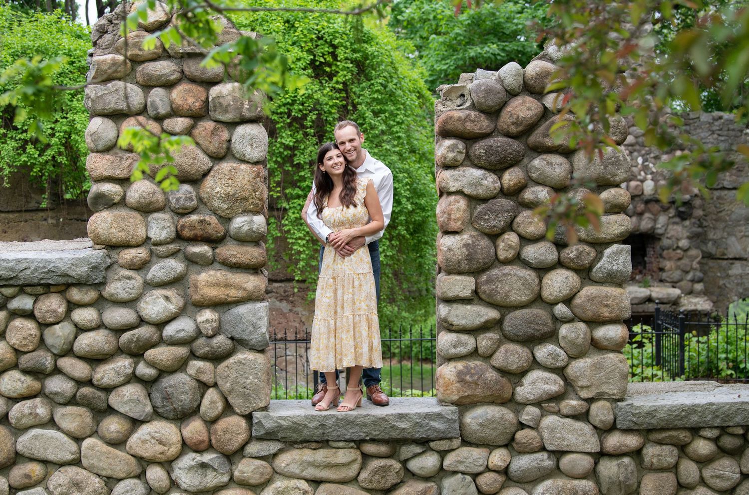 A romantic couple embraces near a rustic stone wall in a lush garden setting with vibrant green foliage.