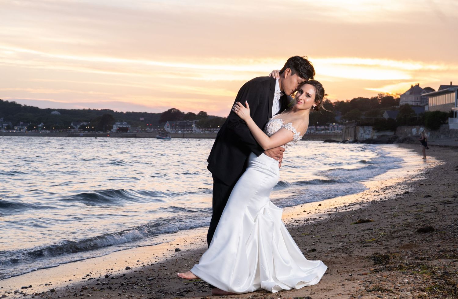 A couple shares a romantic kiss on a sandy beach at sunset with waves gently rolling onto the shore in the background.
