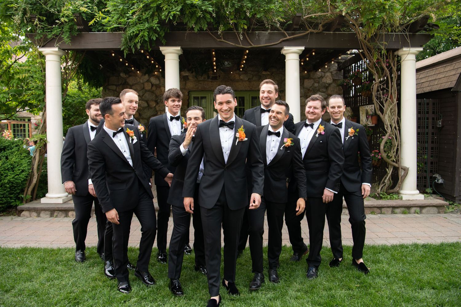 Groom and groomsmen in black tuxedos with orange boutonnieres posing outdoors near white columns and lush greenery.