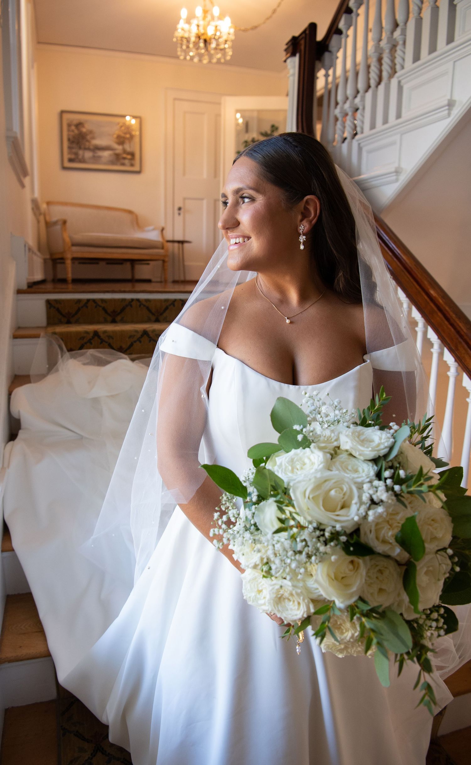 A bride in an elegant off-shoulder white gown holds a bouquet of white roses and greenery while standing by a staircase.