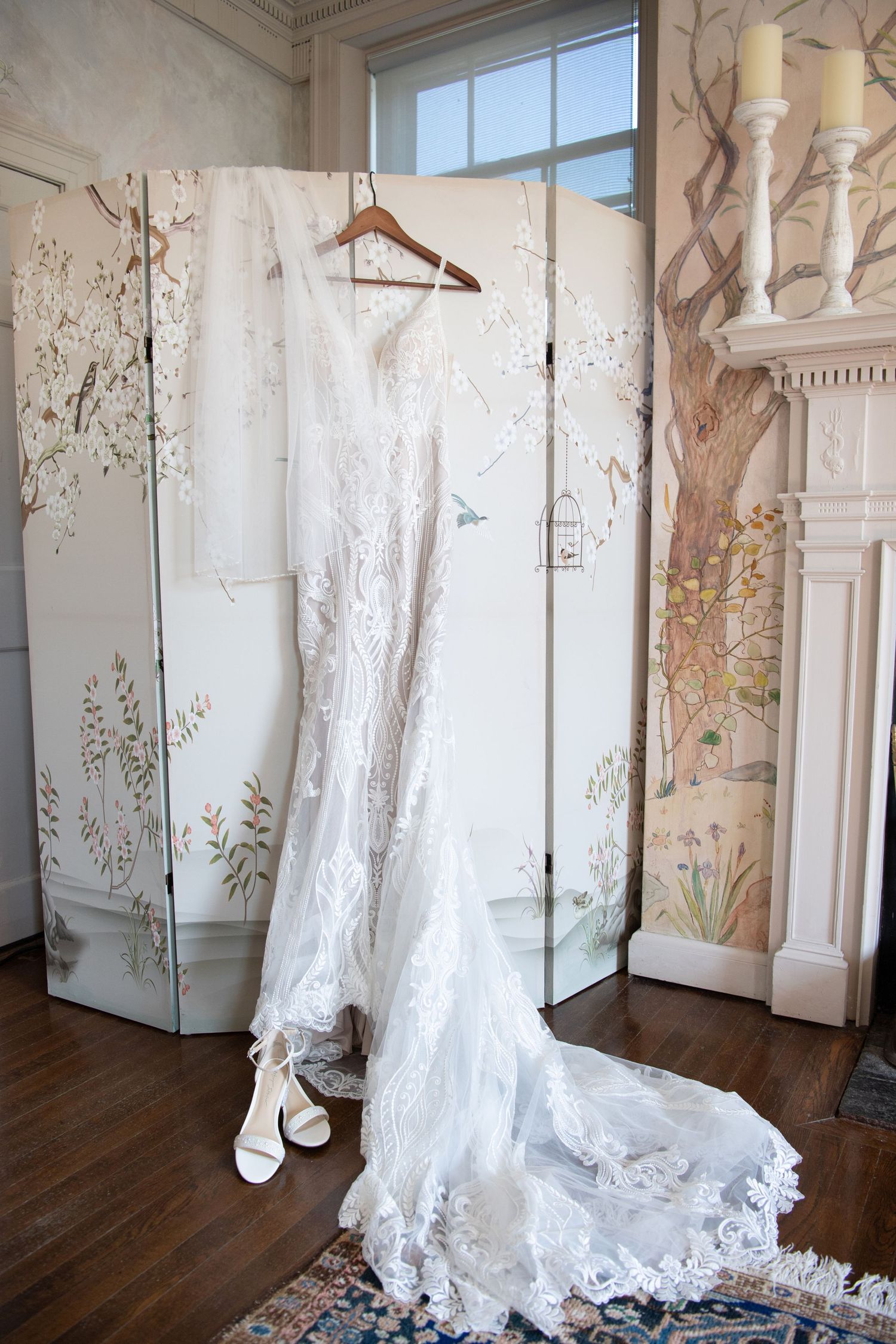 Elegant white lace wedding gown with long train displayed on hanger before floral painted room divider screen.