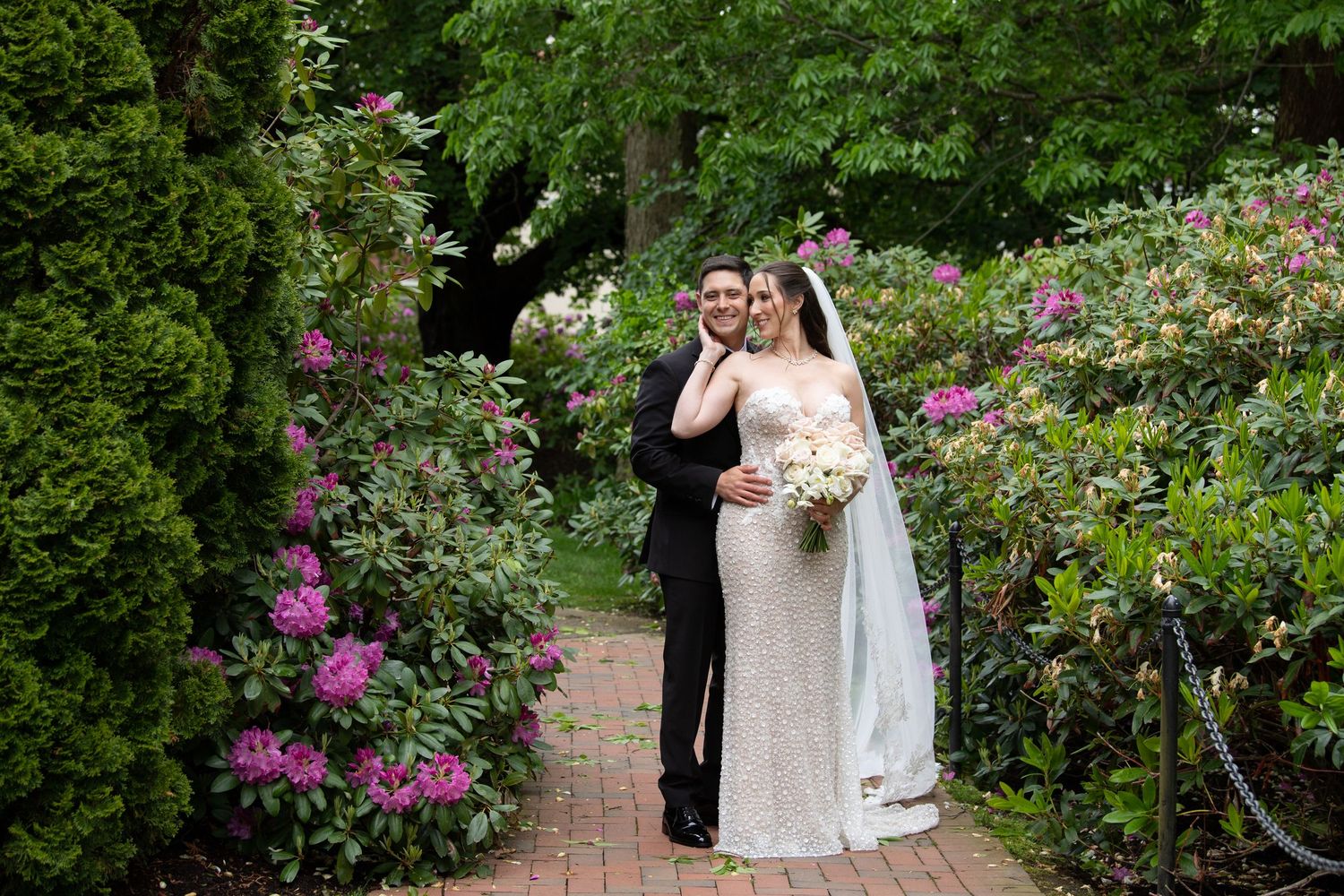 Bride and groom posing on a brick path surrounded by lush greenery and pink rhododendrons in a garden wedding photo.