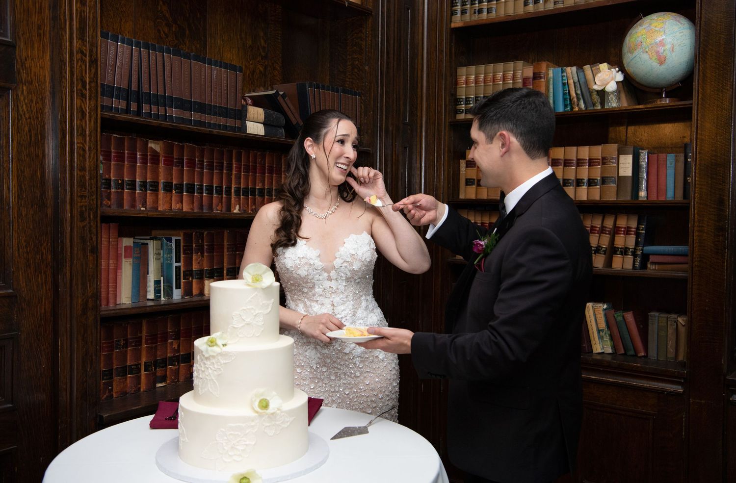 Bride and groom cutting a white three-tier wedding cake in an elegant library setting with bookshelves.