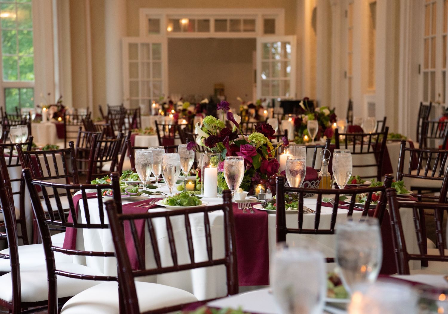 Elegant wedding reception hall with burgundy linens, dark Chiavari chairs, floral centerpieces, and candlelit round tables.